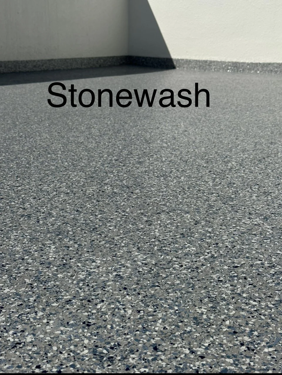 Close-up of a speckled gray textured floor with a corner of a wall and the word 'Stonewash' overlaid.