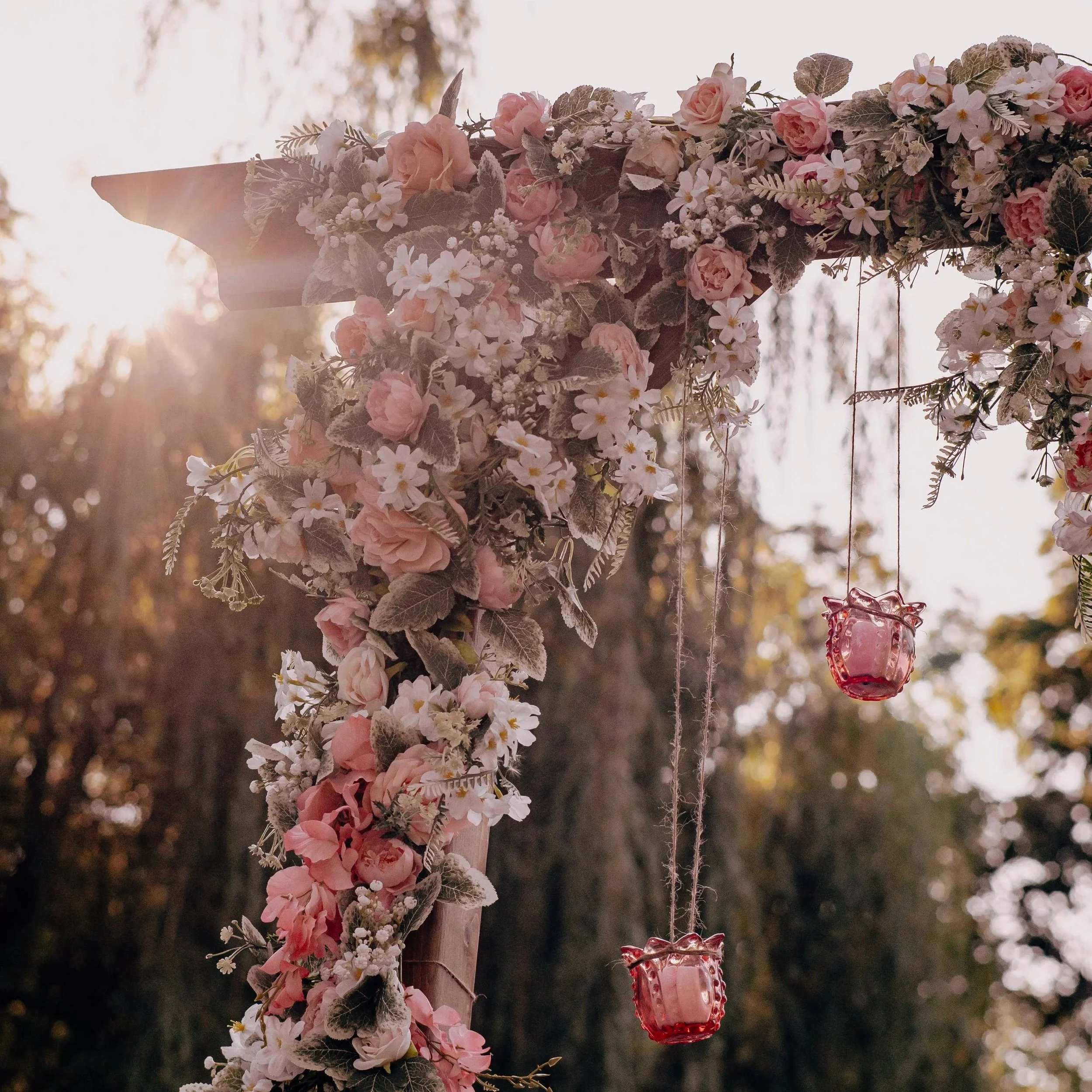 A floral wedding arch decorated with pink and white flowers and green foliage, with hanging pink glass lanterns, outdoors during sunset.