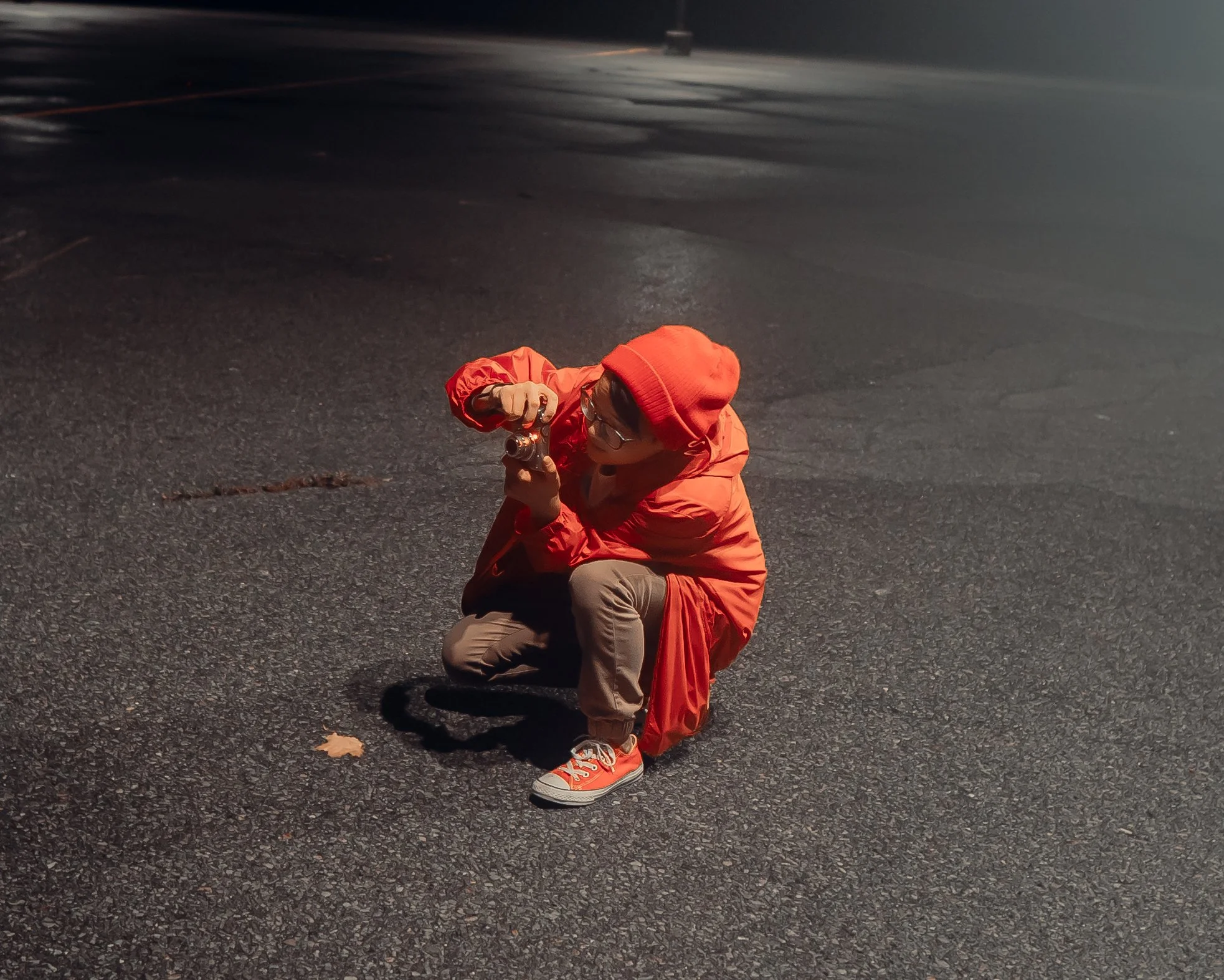 A person in an red jacket, red beanie, and glasses squatting on a wet, dark asphalt surface while taking a photograph with a camera.