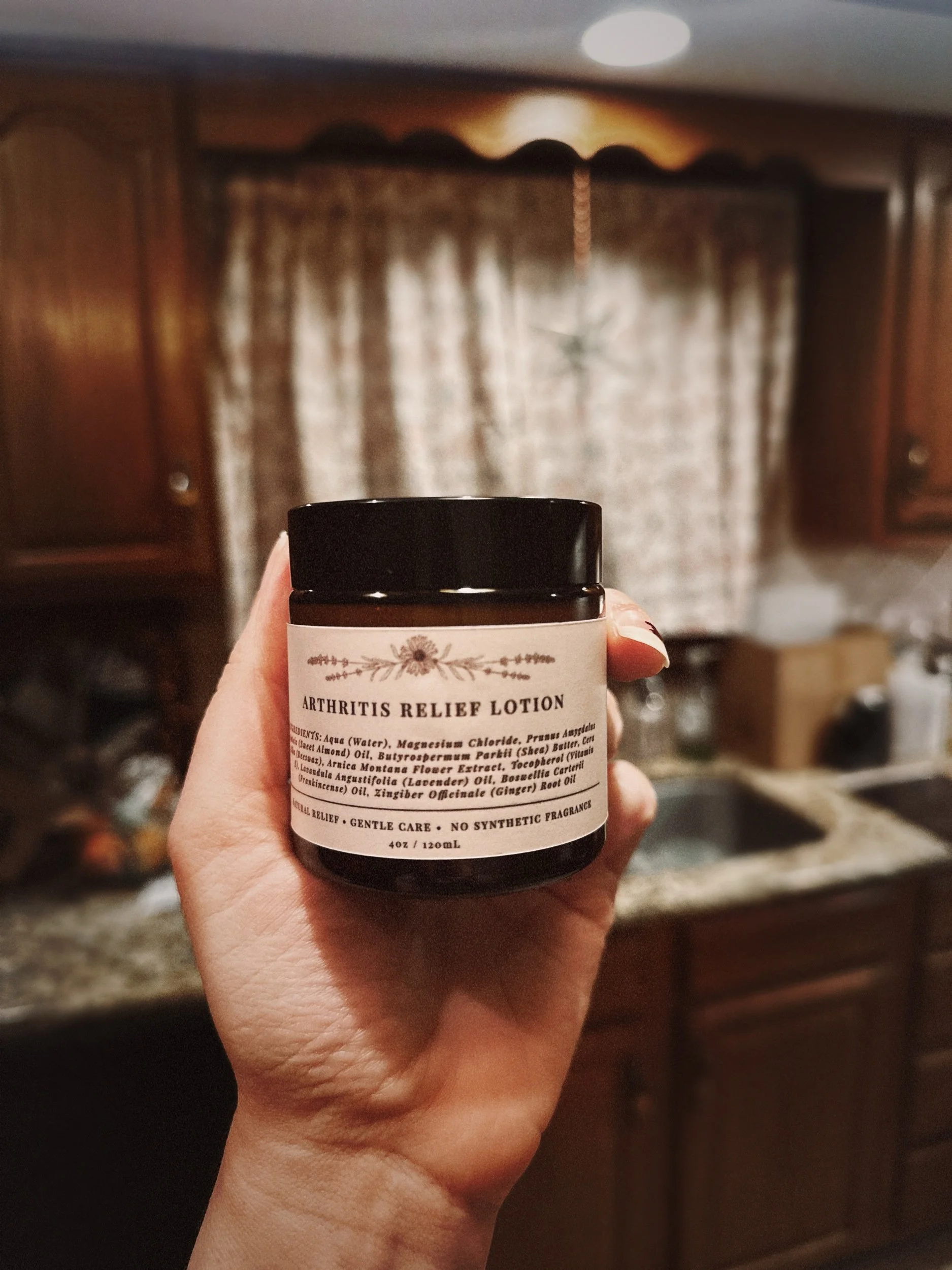 A hand holding a brown jar labeled 'Arthritis Relief Lotion' in a kitchen setting with wooden cabinets, a window with curtains, and a star-shaped decoration hanging in the background.