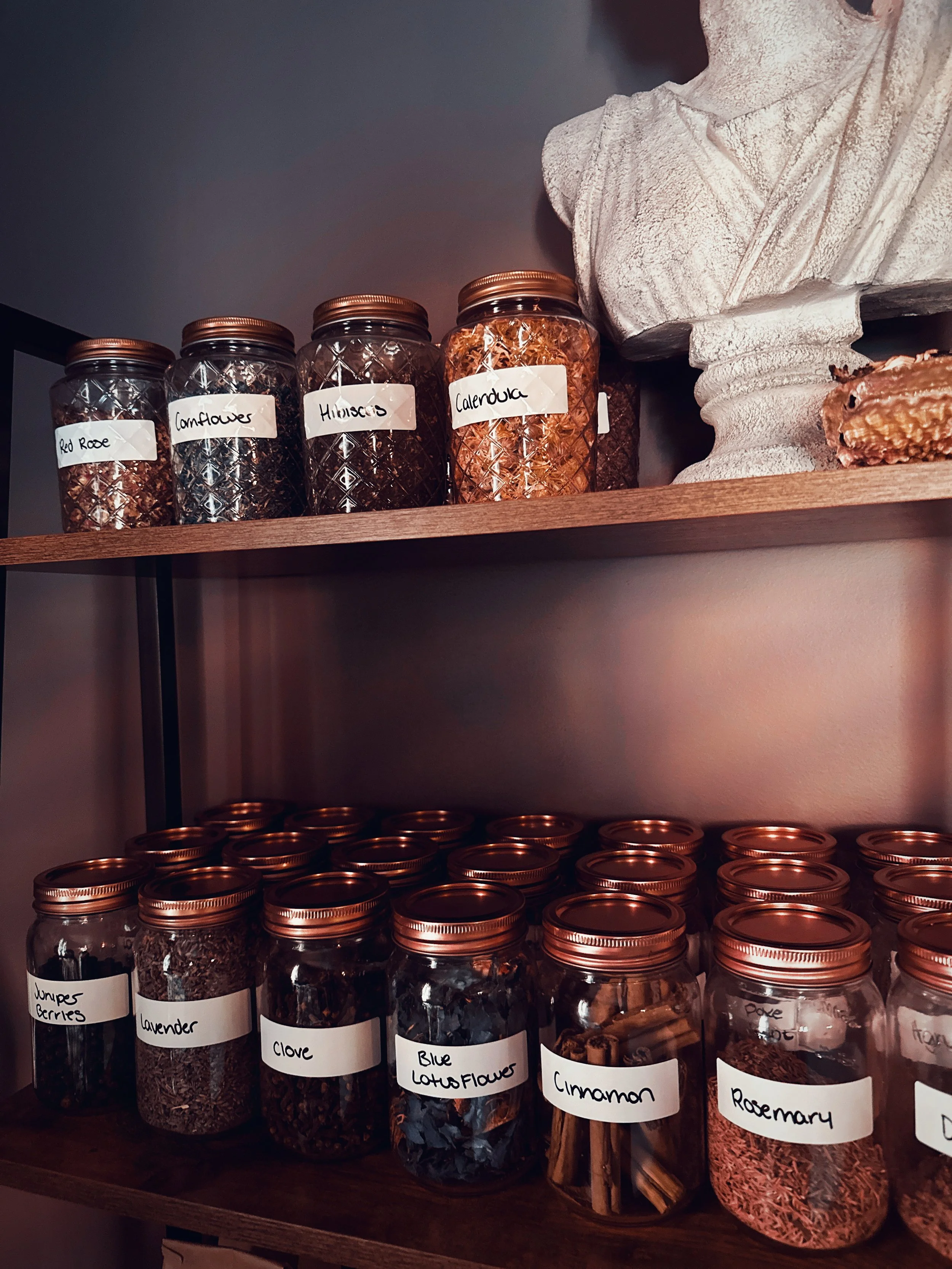 Wooden shelves with glass jars containing dried herbs and spices labeled with names such as red rose, coneflowers, hibiscus, calendula, juniper berries, lavender, clove, blue lotus flower, cinnamon, rosemary, and dandelion.