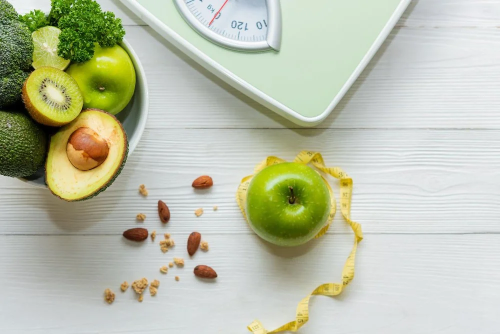 Healthy green foods like avocado, kiwi, and apple with a scale and measuring tape, symbolizing the role of hormones in metabolism, weight management, thyroid function, insulin balance, and cortisol regulation.