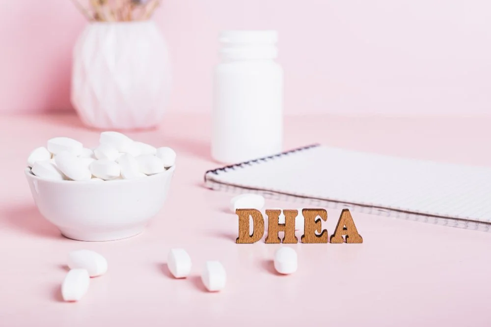 White tablets in a ceramic bowl and scattered on a pink table next to a notebook and a bottle, with wooden letters spelling 'DHEA' — symbolizing hormone supplementation for adrenal and hormonal health in women.