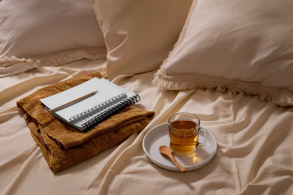 Notebook, pencil, and cup of herbal tea on a cozy bed—symbolizing relaxation, journaling, and calming bedtime rituals that support progesterone and restful sleep.