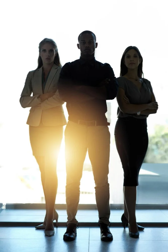 Three business professionals standing with arms crossed in front of a bright window, backlit with sunlight.