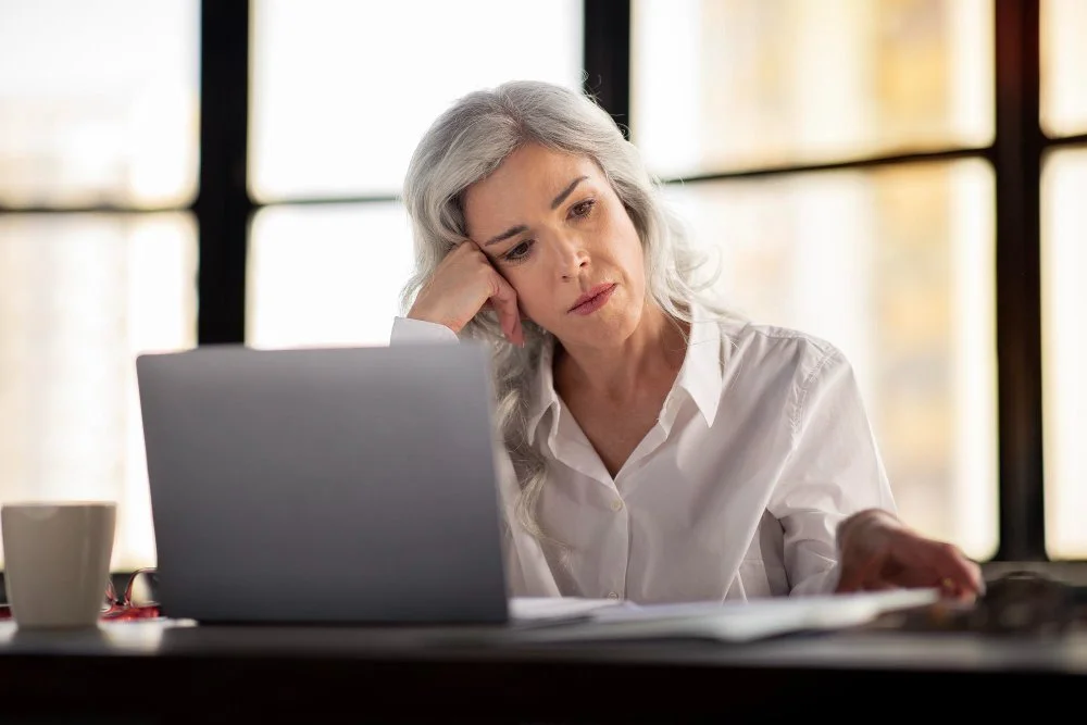 A thoughtful older woman with long gray hair sitting at a desk, looking fatigued while working on a laptop, with papers and a coffee mug nearby. This image reflects the theme of fatigue, hormonal imbalance, and low testosterone in women.
