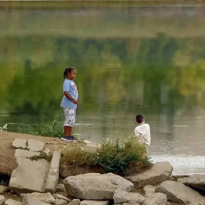 Two children discovering the Water Element while standing beside a calm lake on a rocky shore