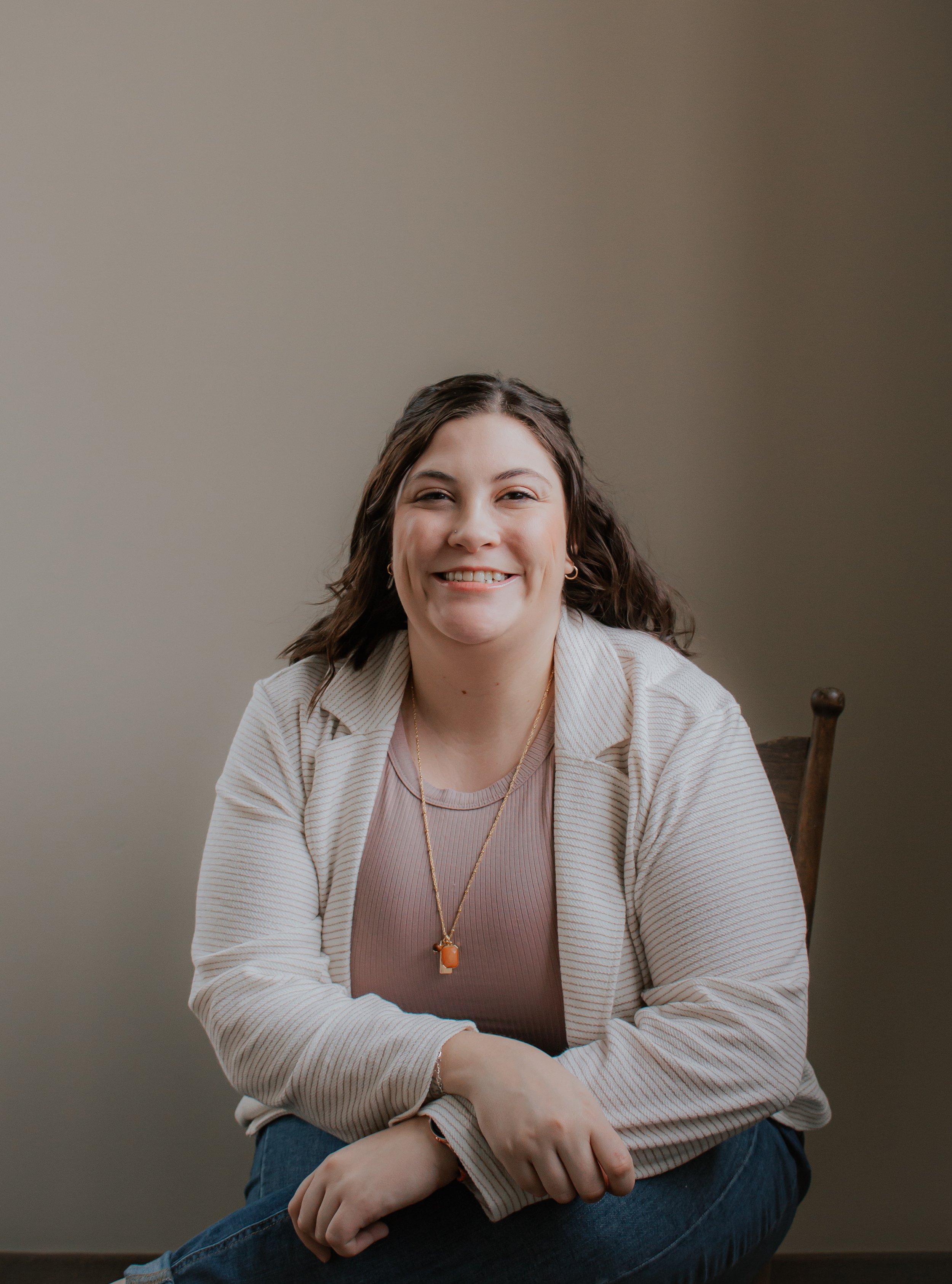A woman with dark wavy hair smiling, sitting on a wooden chair with neutral background, wearing a beige blazer, pink top, and a gold necklace.