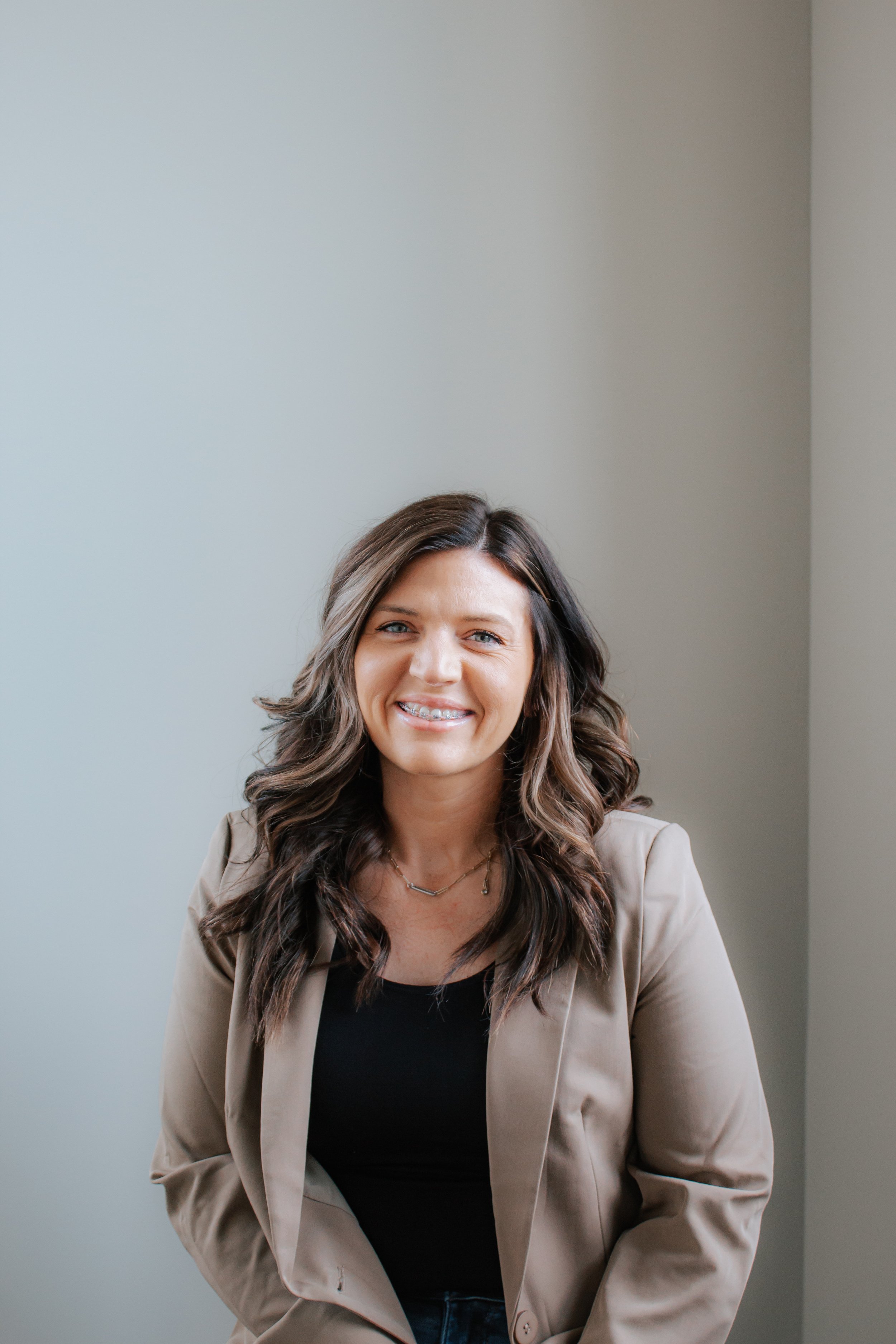 Smiling woman with wavy brown hair, wearing a beige blazer and black top, standing against a plain light-colored wall.