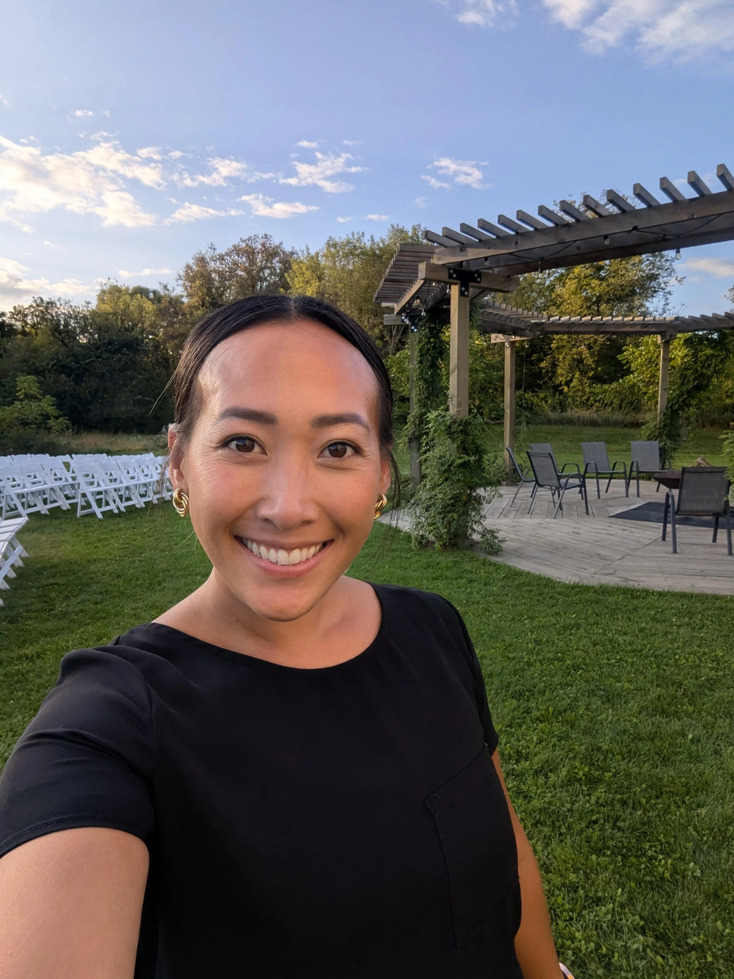 Smiling woman taking selfie outdoors in a grassy area during the daytime, with a wooden pergola and outdoor chairs in the background, trees, and a partly cloudy sky.