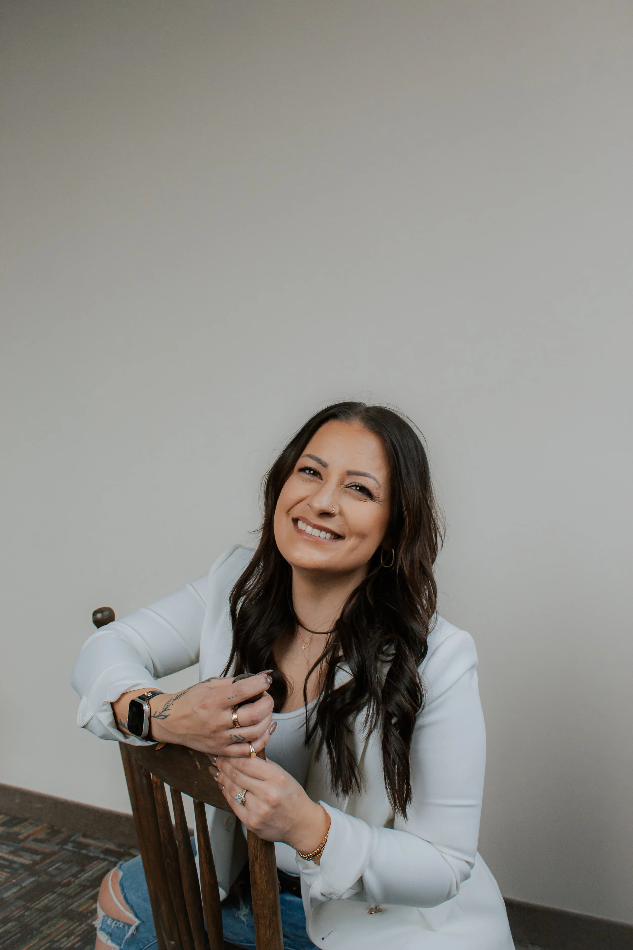 A woman with long dark hair smiling and sitting on a wooden chair against a plain wall.