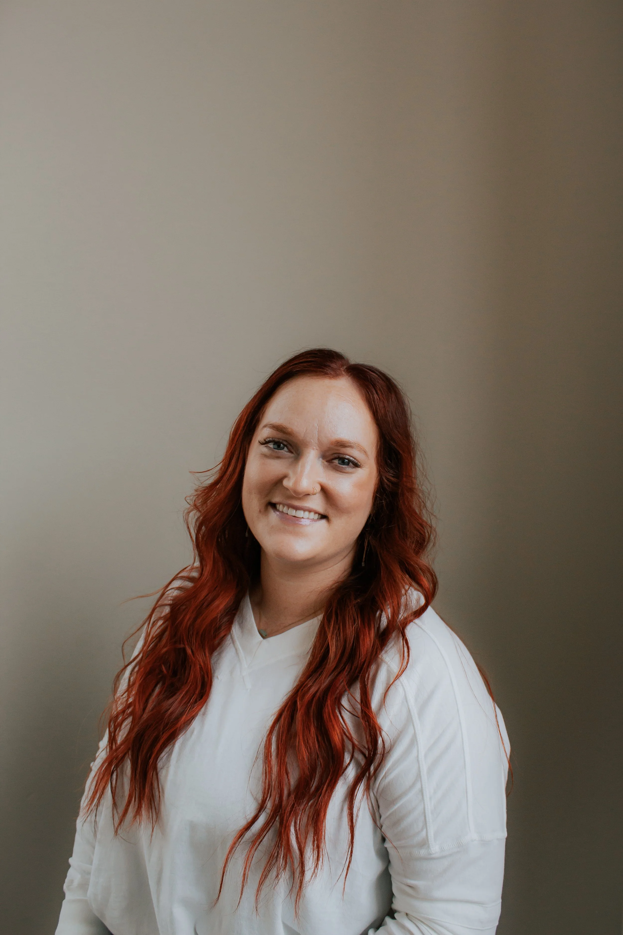 A woman with long, wavy red hair and a nose ring, smiling and wearing a white shirt, standing against a plain beige wall.