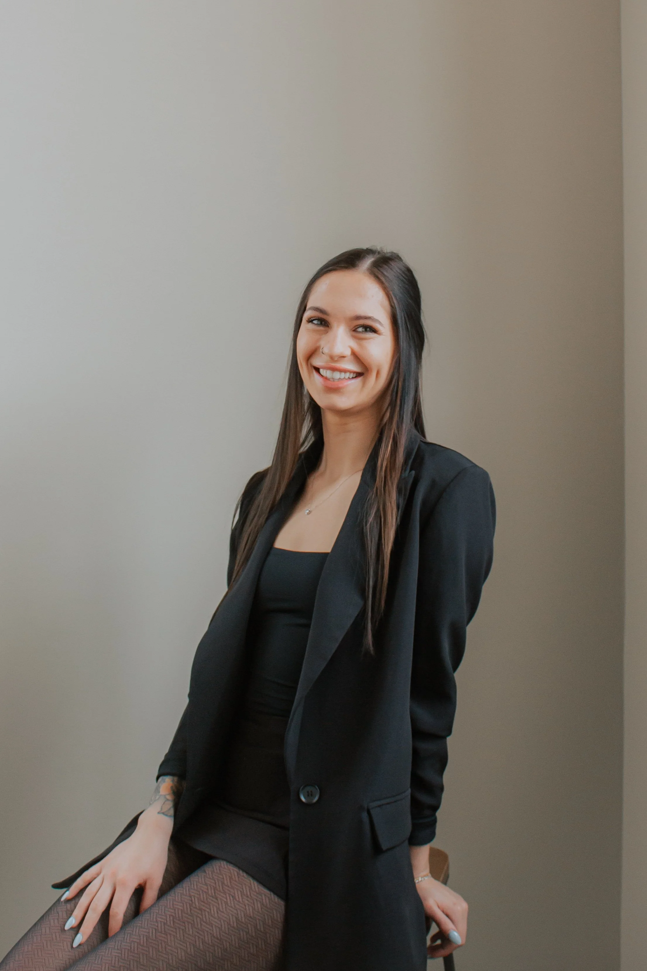 A woman with long dark hair sitting on a stool against a plain wall, smiling at the camera.