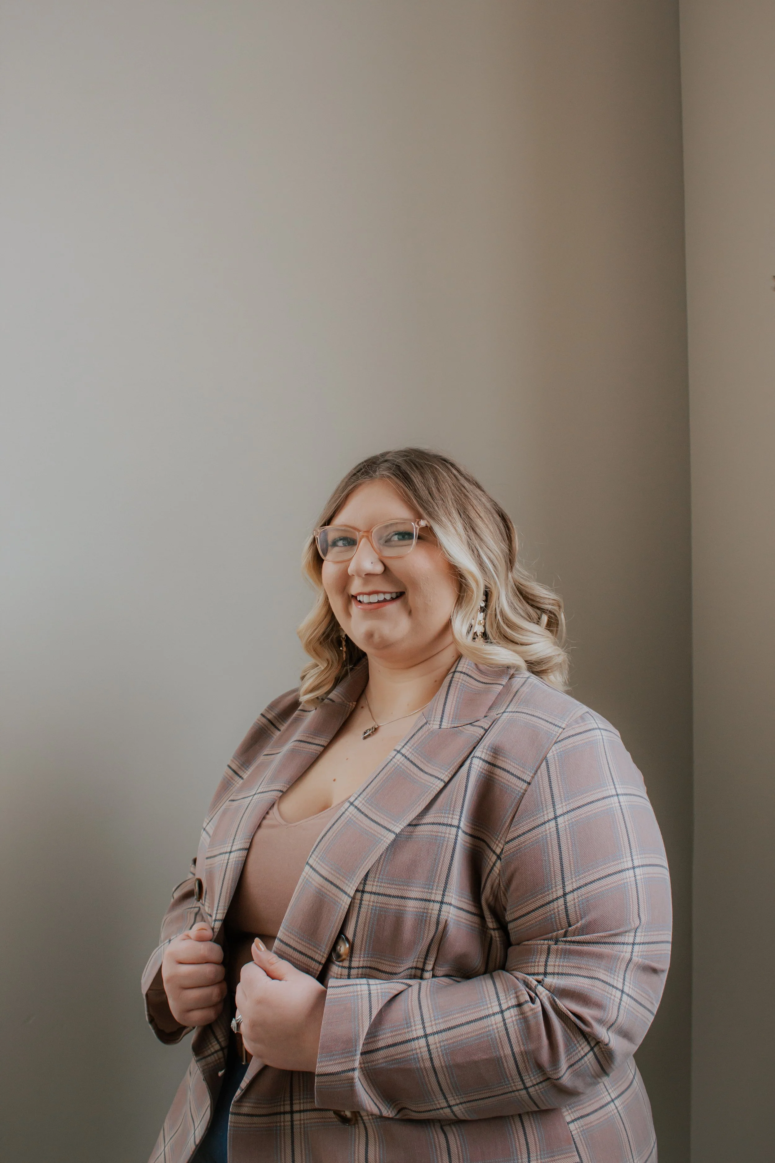 A woman with blonde, wavy hair, glasses, and a friendly smile standing against a plain, light-colored wall. She is wearing a plaid blazer over a beige top and holding the lapels of her blazer.