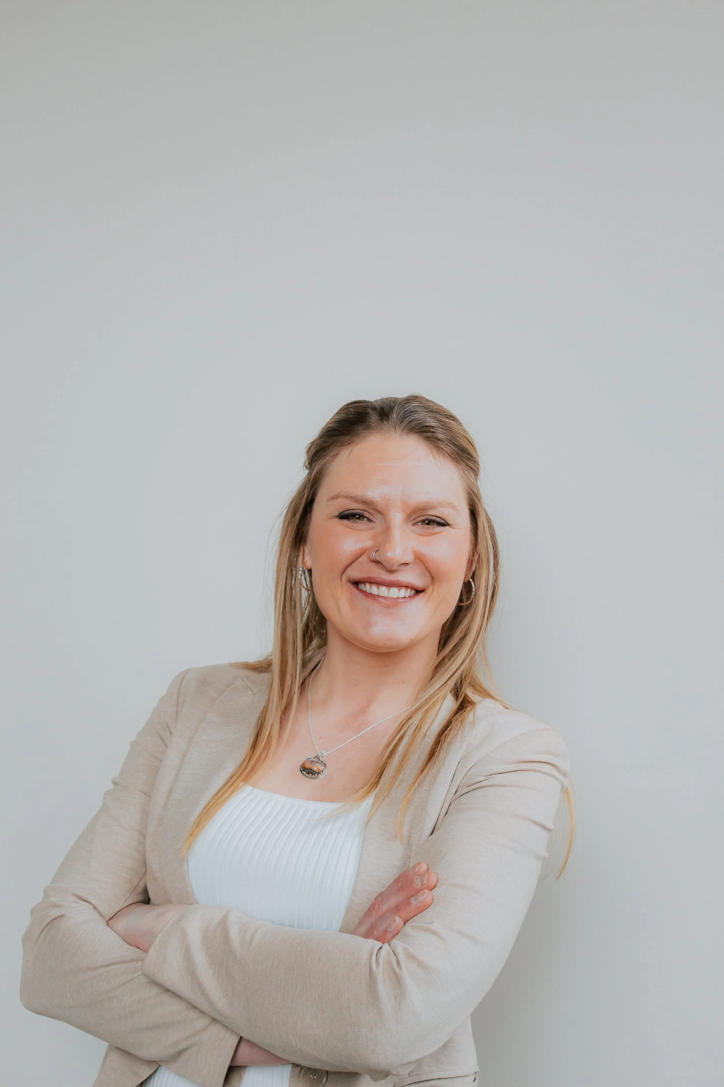 A woman with long blonde hair, arms crossed, smiling, wearing a beige blazer over a white top, against a plain white background.