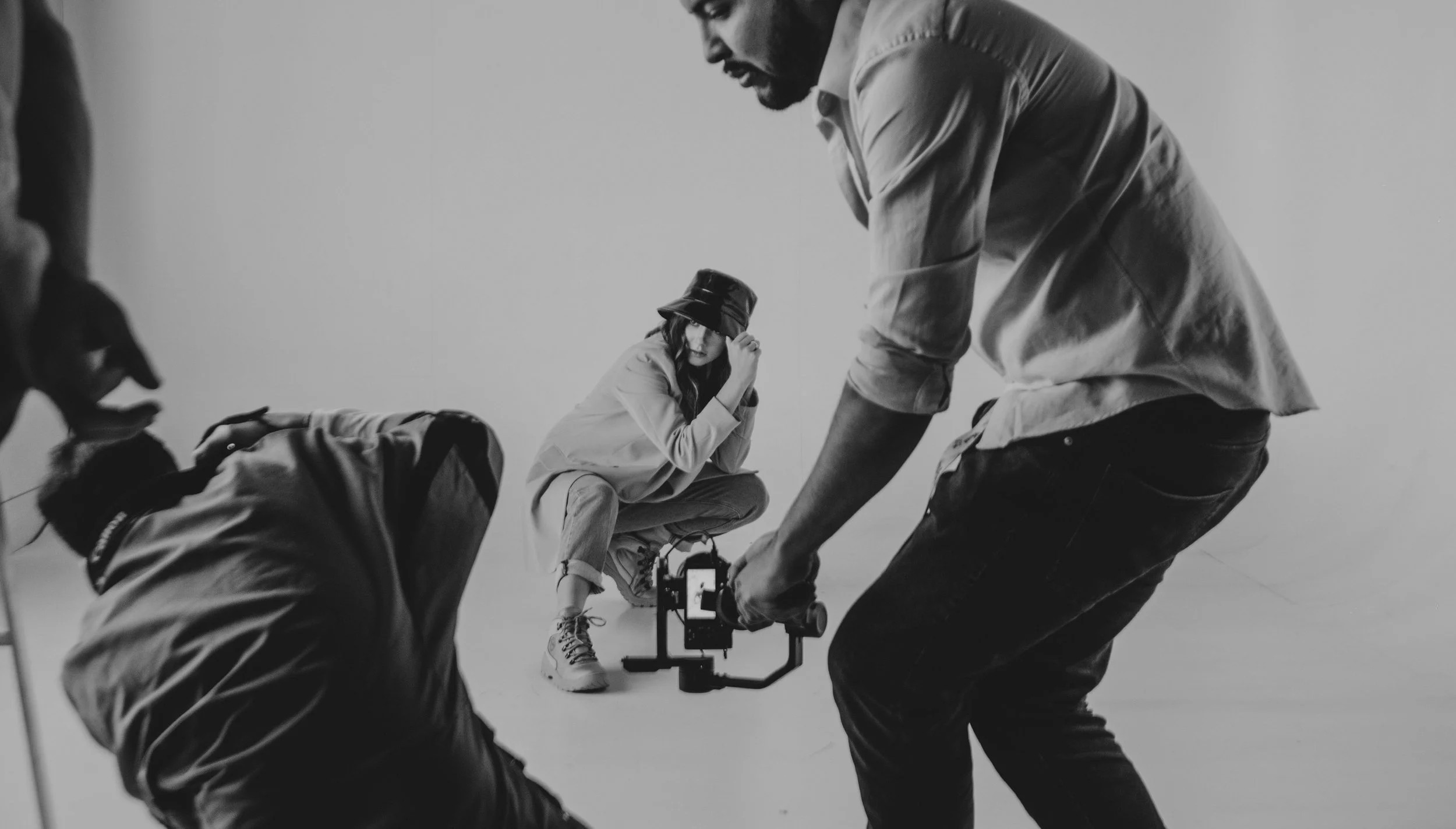 Black and white photo of a photoshoot with a woman wearing a bucket hat and sneakers sitting on a stool, while three crew members assist her and set up equipment.