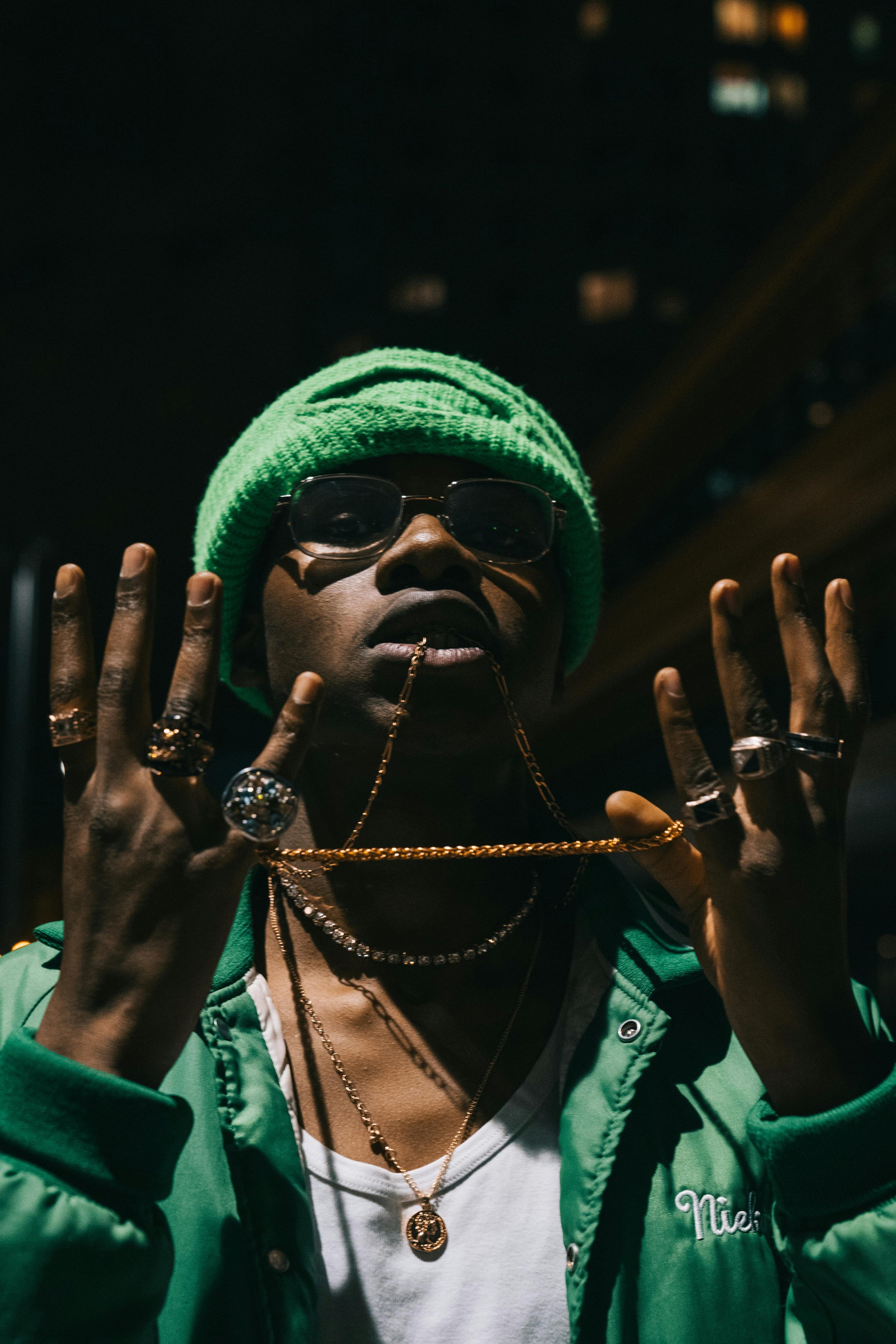 A person wearing a bright green knitted beanie, sunglasses, multiple rings, and layered gold chains, dressed in a green jacket and white shirt, making a peace sign with both hands at night in an urban environment.