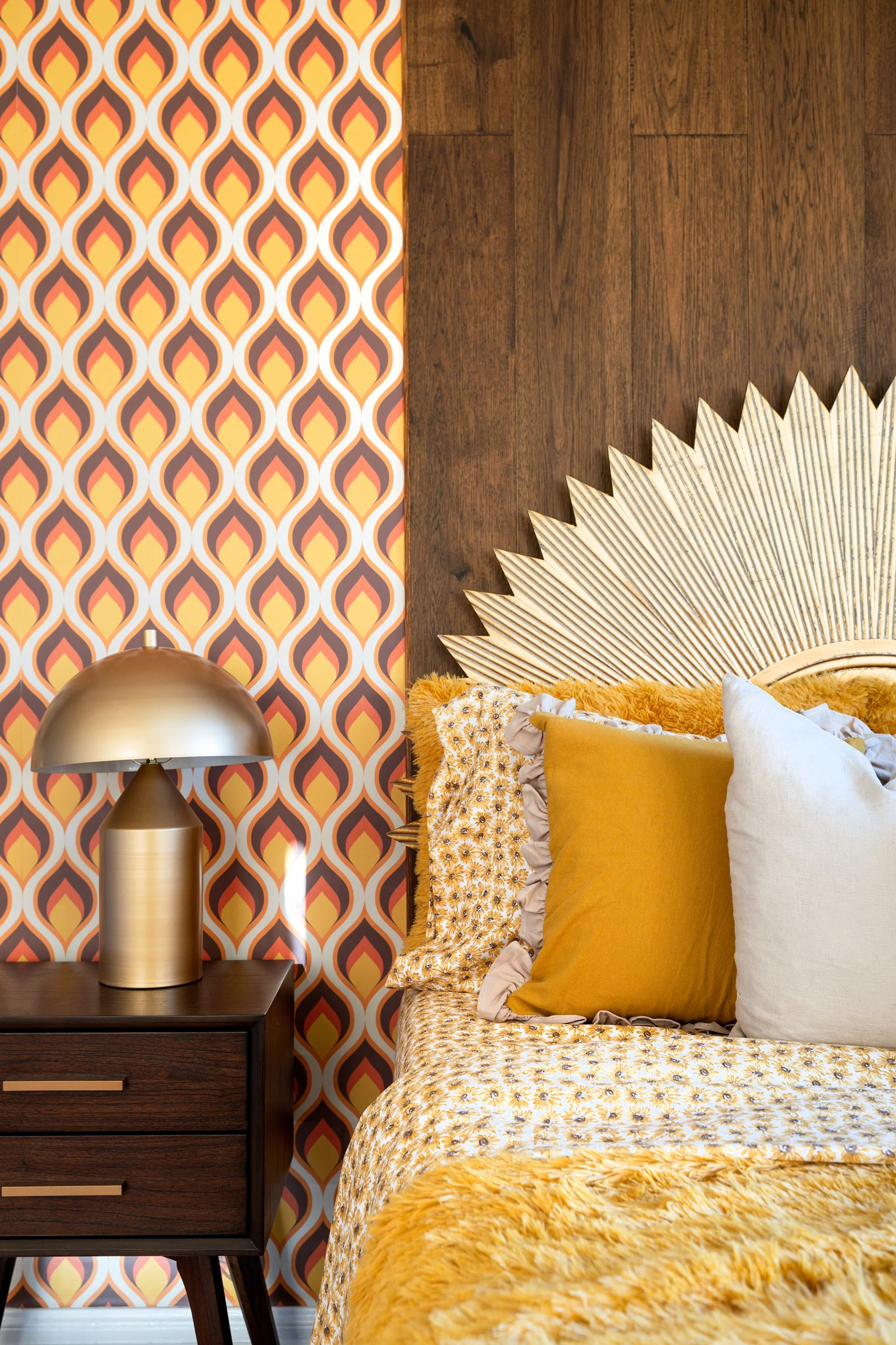 Retro bedroom interior with bold patterned wallpaper, sunburst headboard, mustard and cream pillows, and a vintage-style lamp on a wooden nightstand.
