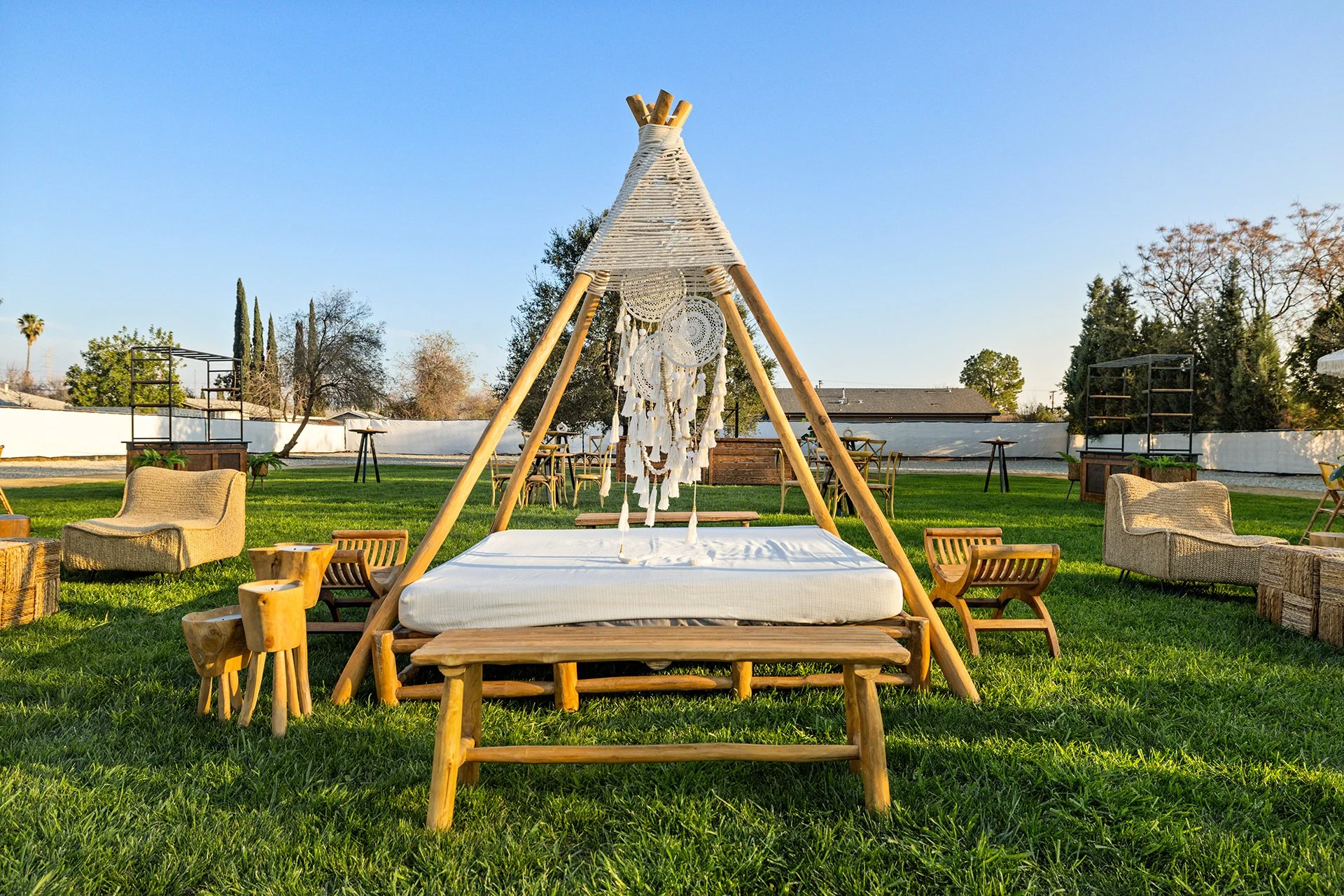 Outdoor seating area with teepee-style canopy, wooden benches, chairs, and tables on a grassy lawn.