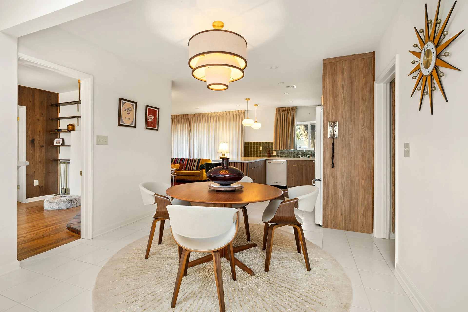 Mid-century modern dining room with a round wooden table, white chairs, and a large pendant light. A sunburst clock is on the wall, and the room connects to a kitchen with wood cabinetry and green tile backsplash.