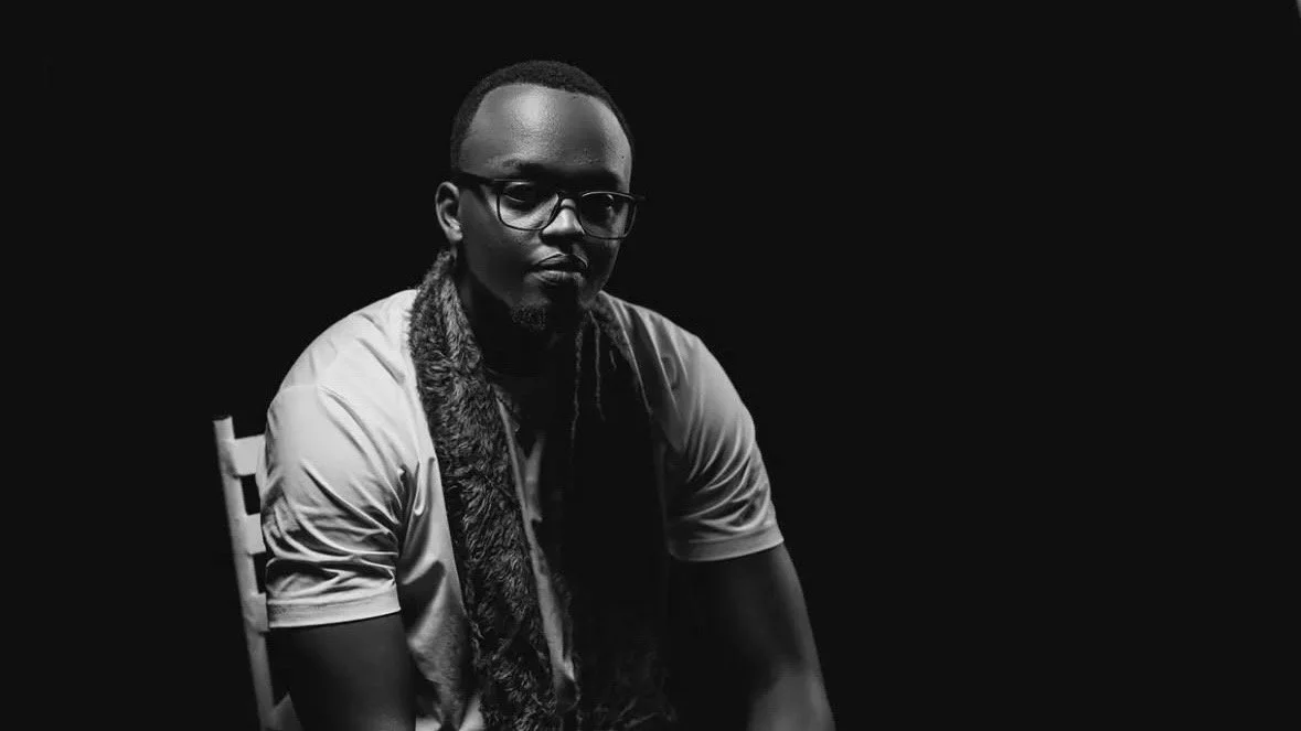Black and white photo of a man with glasses, short hair, and facial hair, sitting on a chair against a dark background, looking serious.