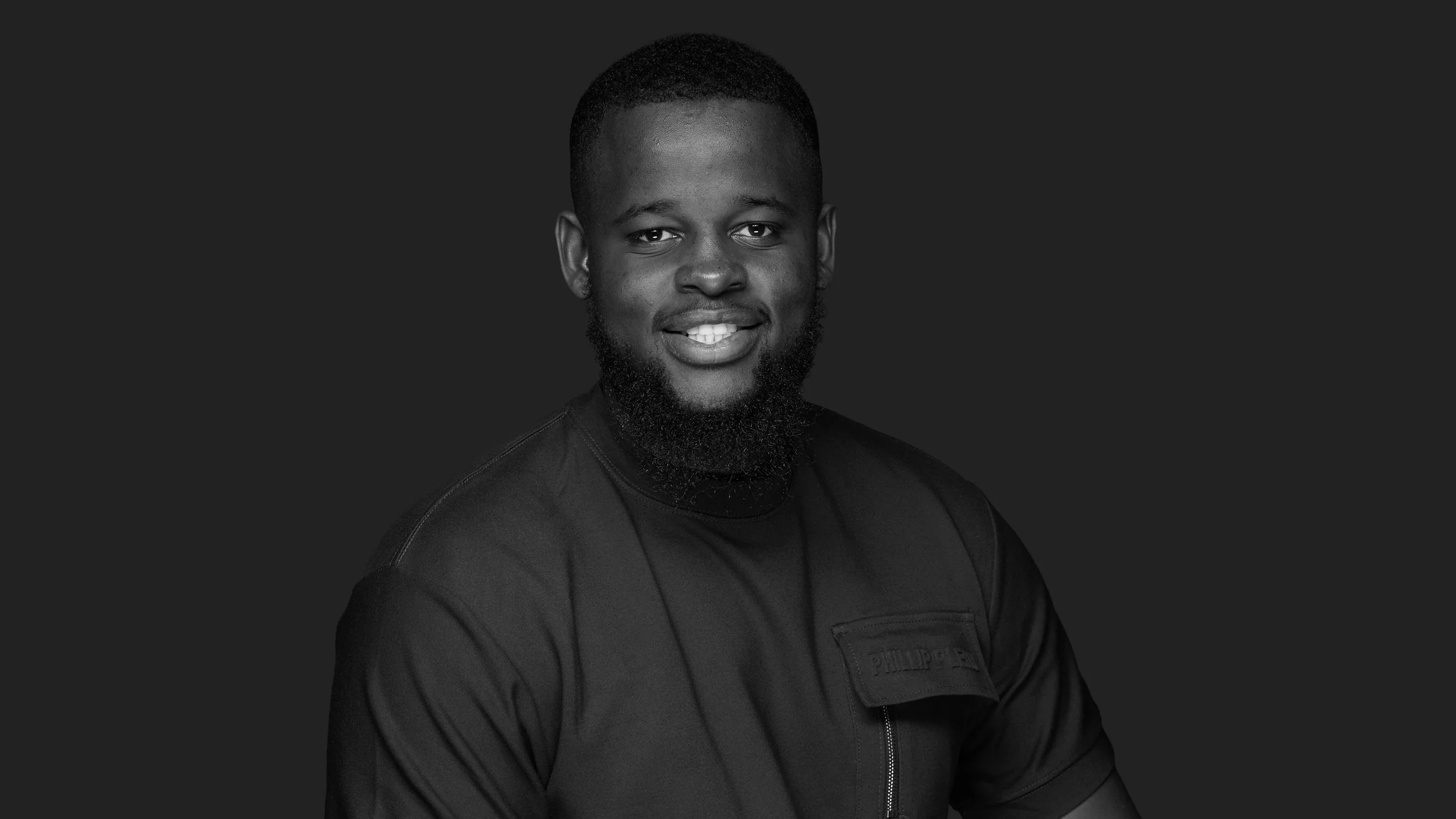 Black and white portrait of a smiling young man with a beard, wearing a jacket, against a dark background.