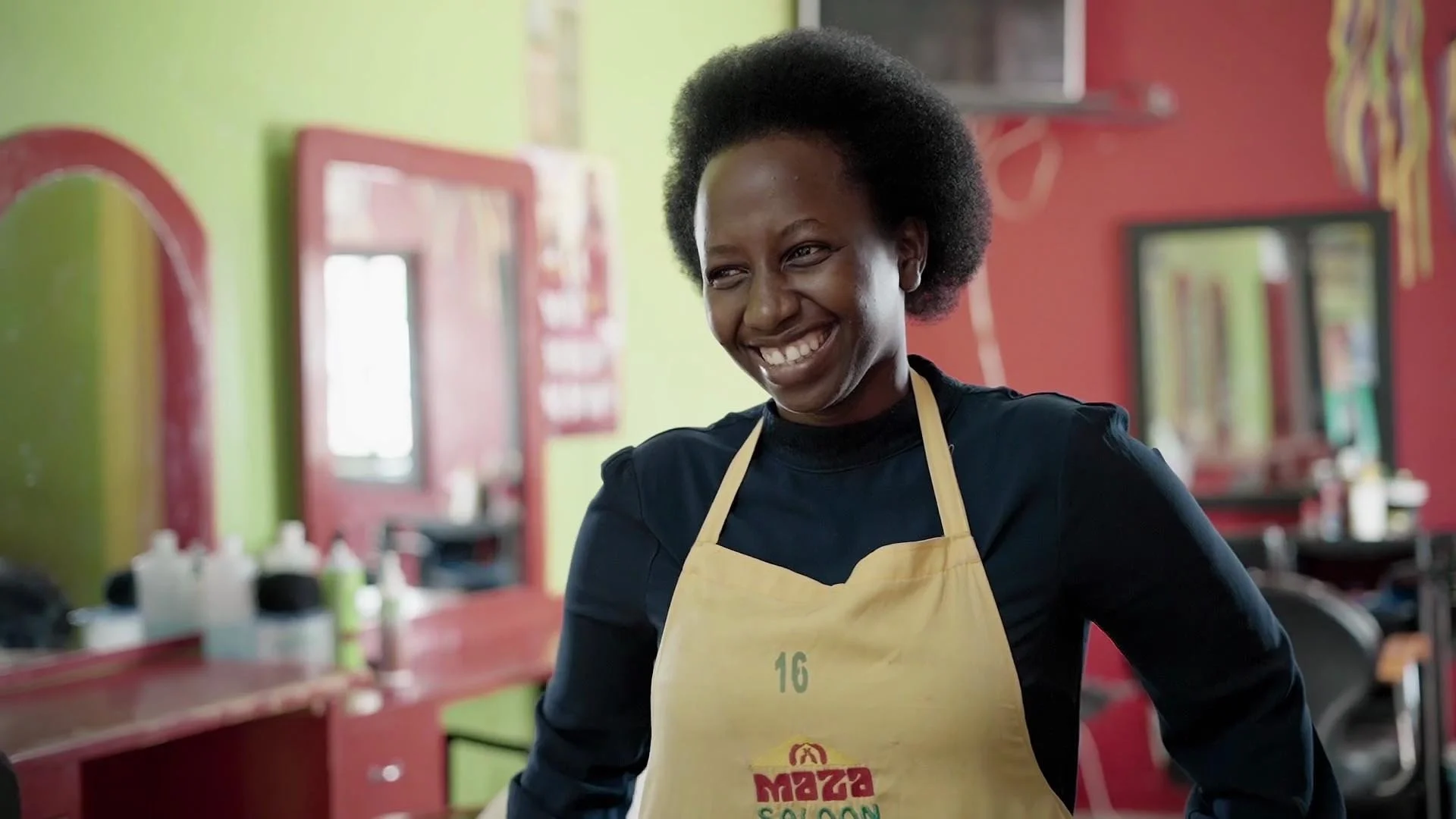 A woman with natural hair smiling in a hair salon wearing a yellow apron.