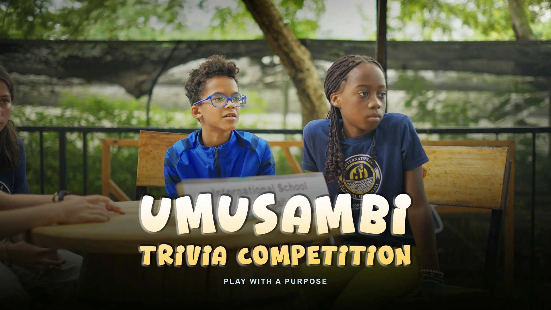 Children participating in a trivia competition outdoors, sitting at a table with a sign that reads 'UMUSAMBI Trivia Competition', with a green, leafy background.