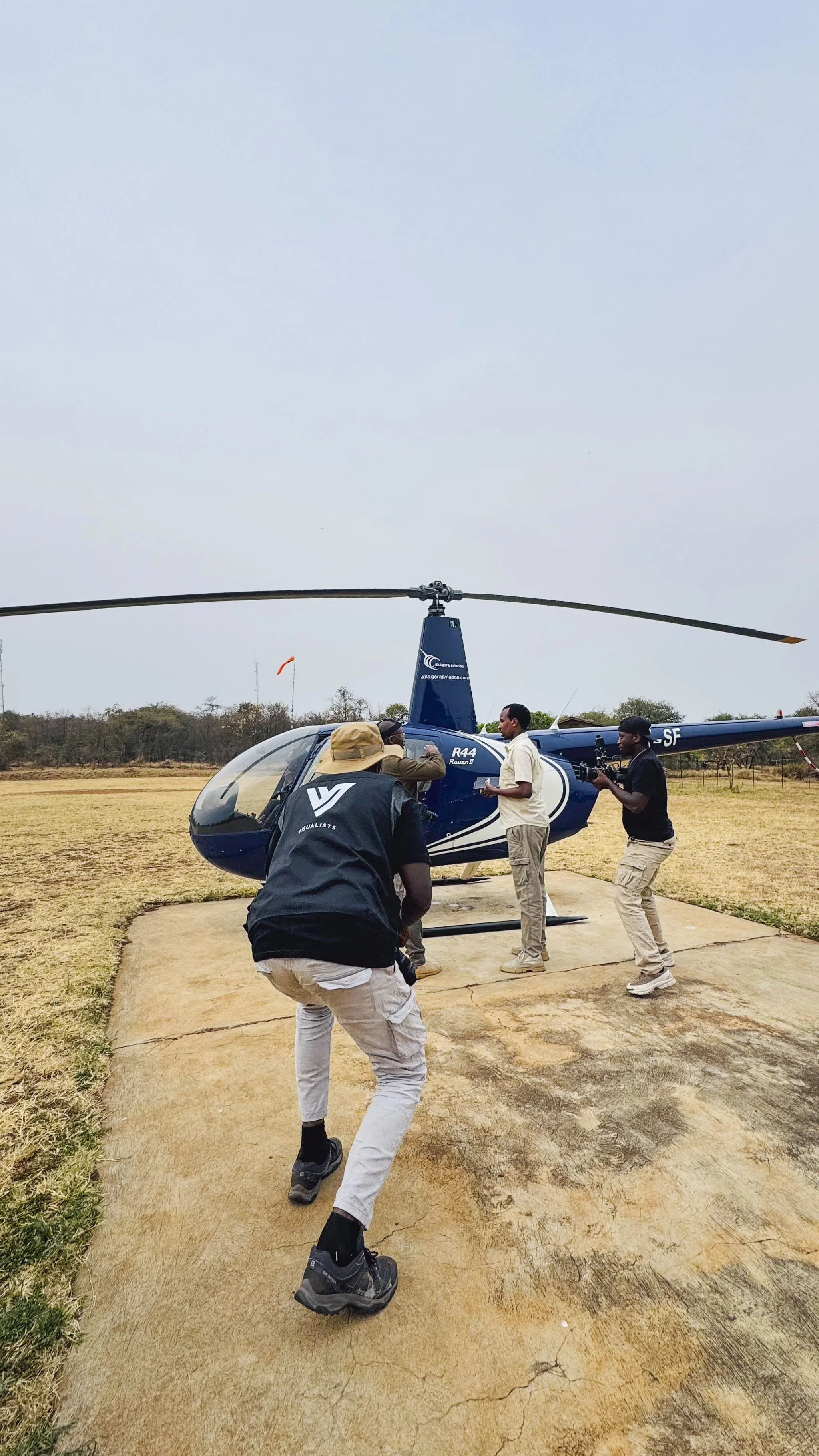 Behind the scene of Visualists Team filming and photographing researching who are about to hop on a blue helicopter on a concrete pad in an open field.