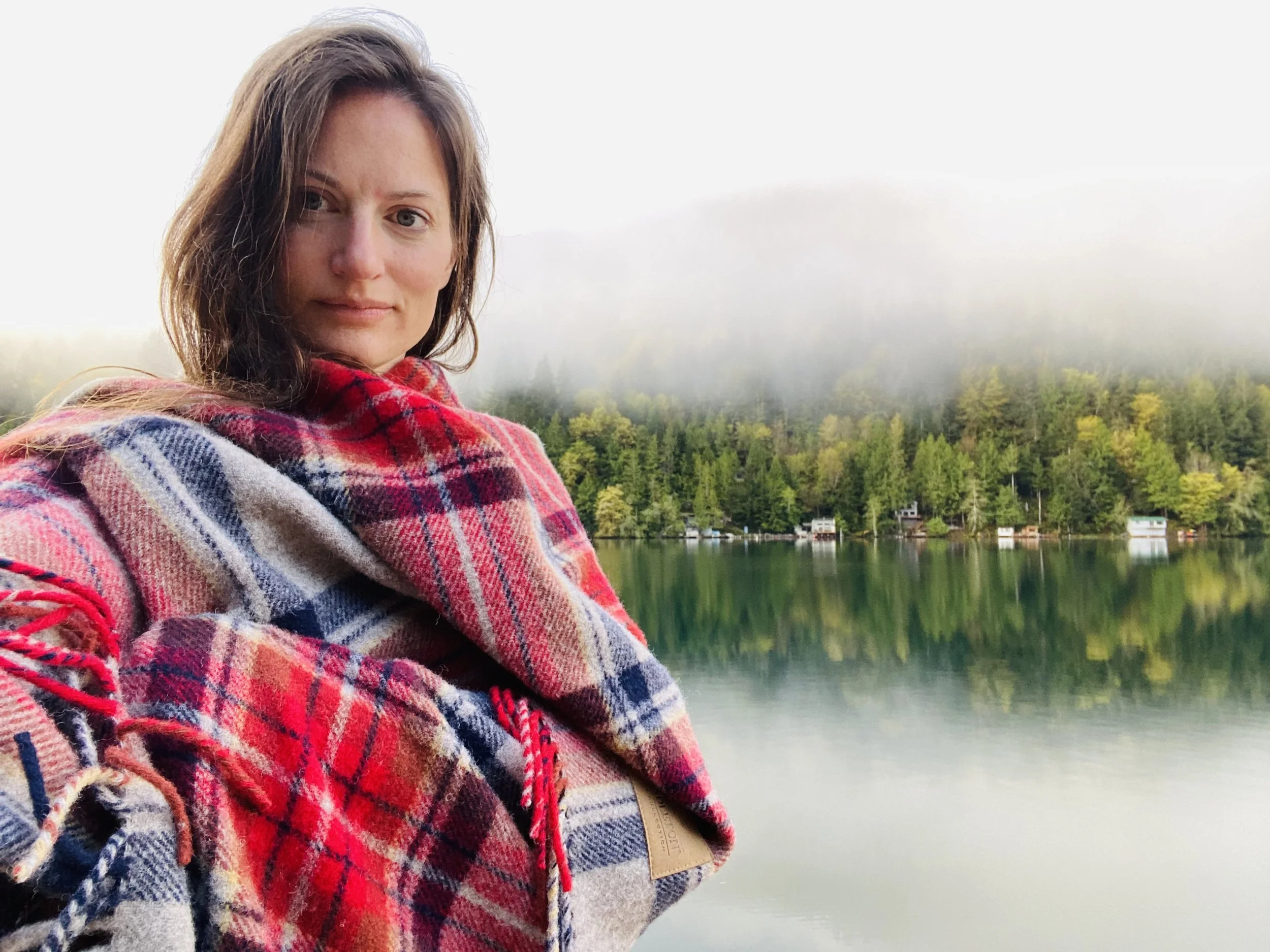 A woman weathering a colorful plaid scarf standing by a body of water with lush green trees in the background, under a foggy or cloudy sky.