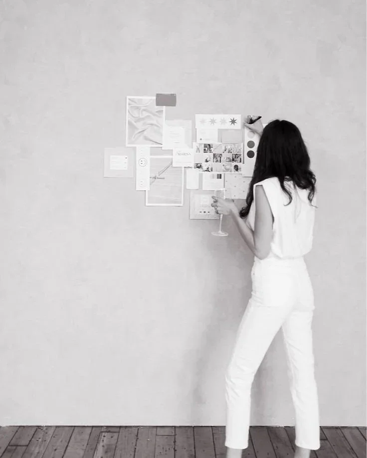 Woman in white pants and sleeveless top standing by a wall decorated with various papers, photos, and color swatches, holding a drink.