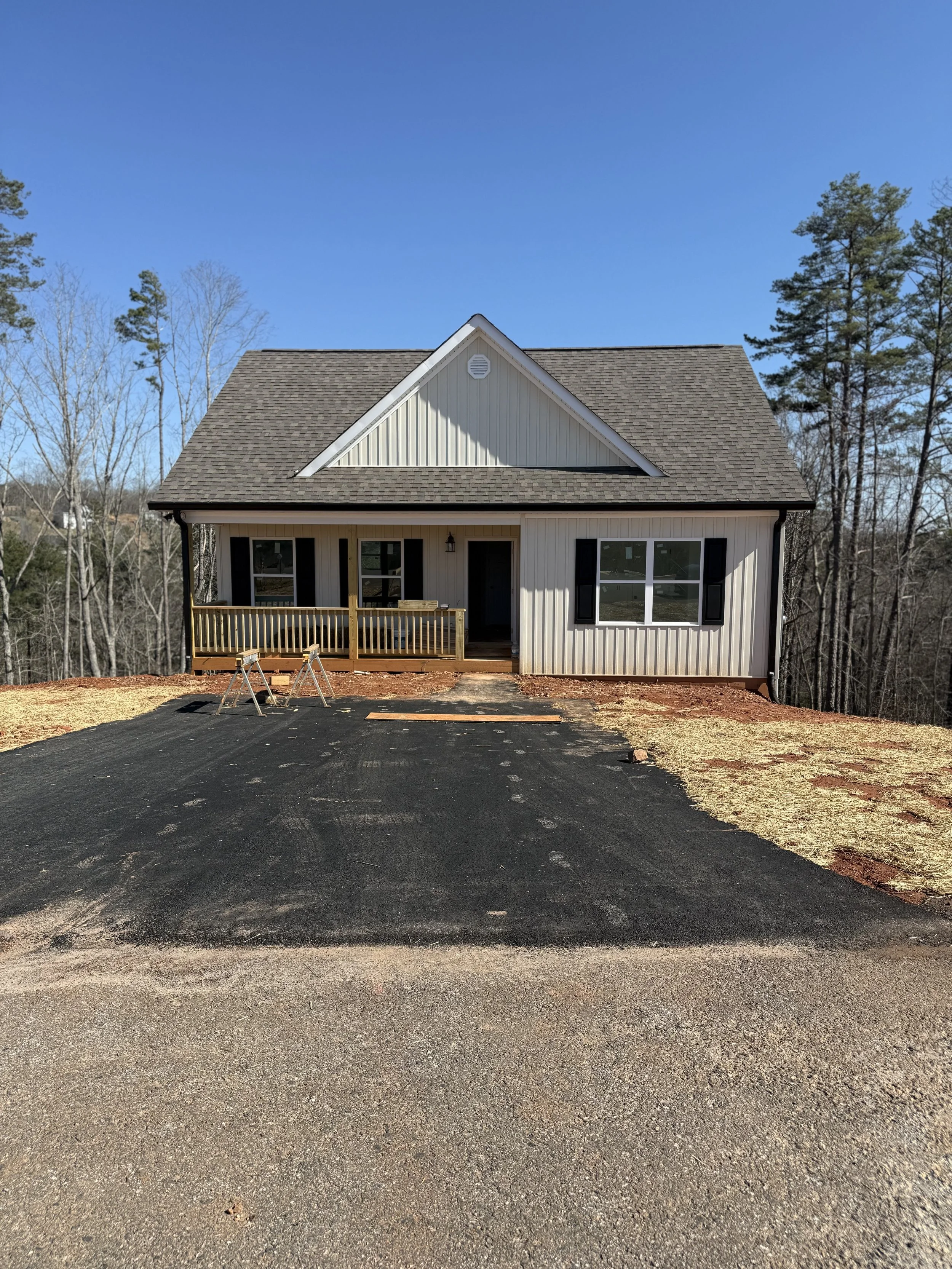 Newly constructed house with a dark asphalt driveway, white siding, and black shutters, on a clear day with a blue sky and trees in the background.