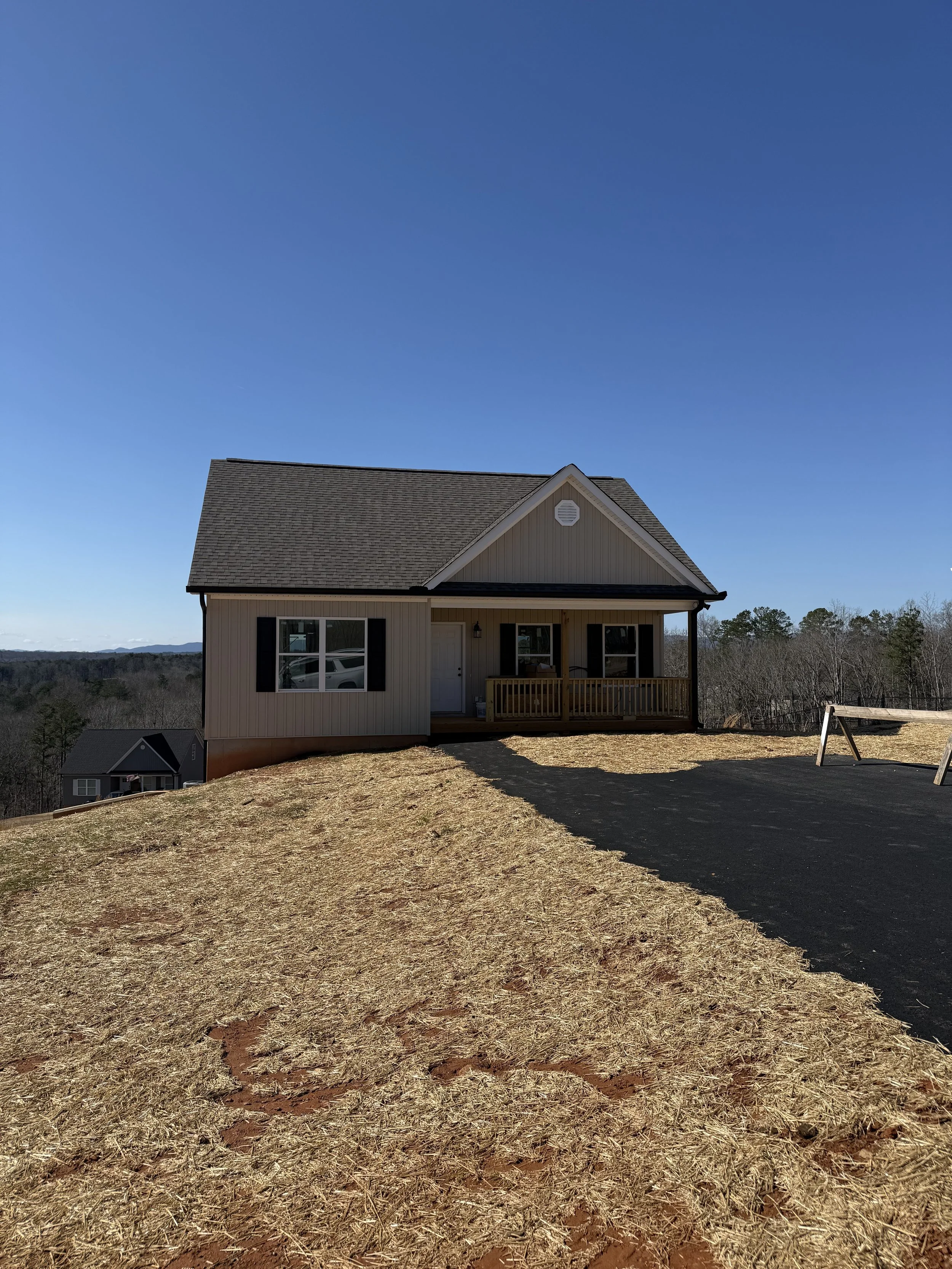 New house on a hill with a paved pathway in front, surrounded by open land and trees, under a clear blue sky.