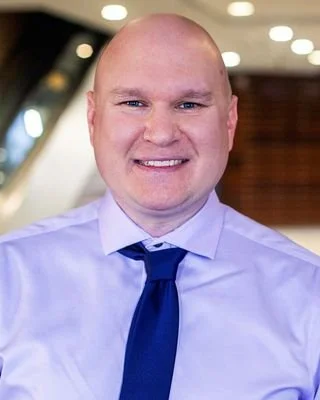 A smiling man dressed in a light blue shirt and a dark blue tie, with a blurred indoor background.