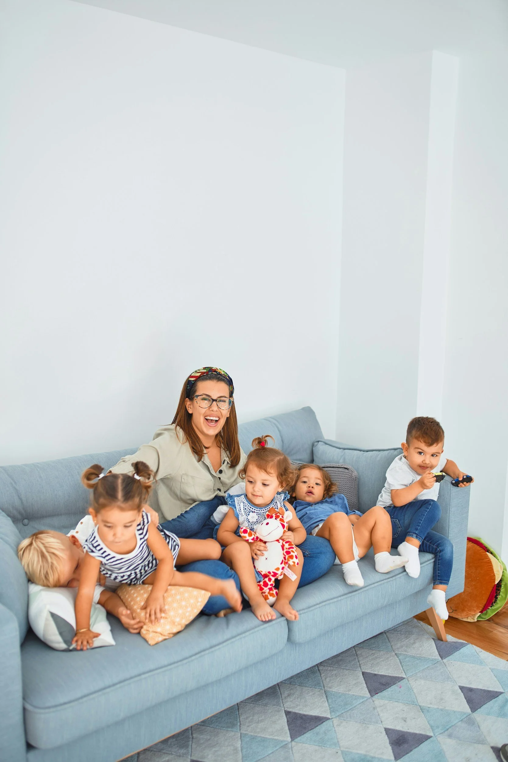 Woman with glasses playing with five children on a blue couch in a living room.