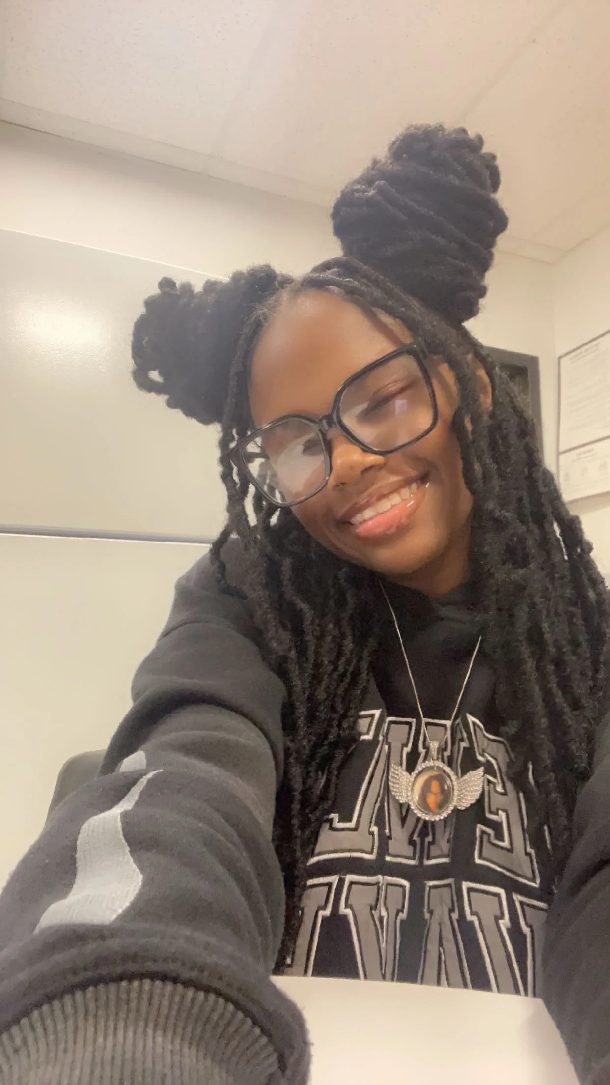 Smiling woman with long dreadlocks styled in big buns, wearing glasses, a black hoodie, and a necklace, taking a selfie indoors.