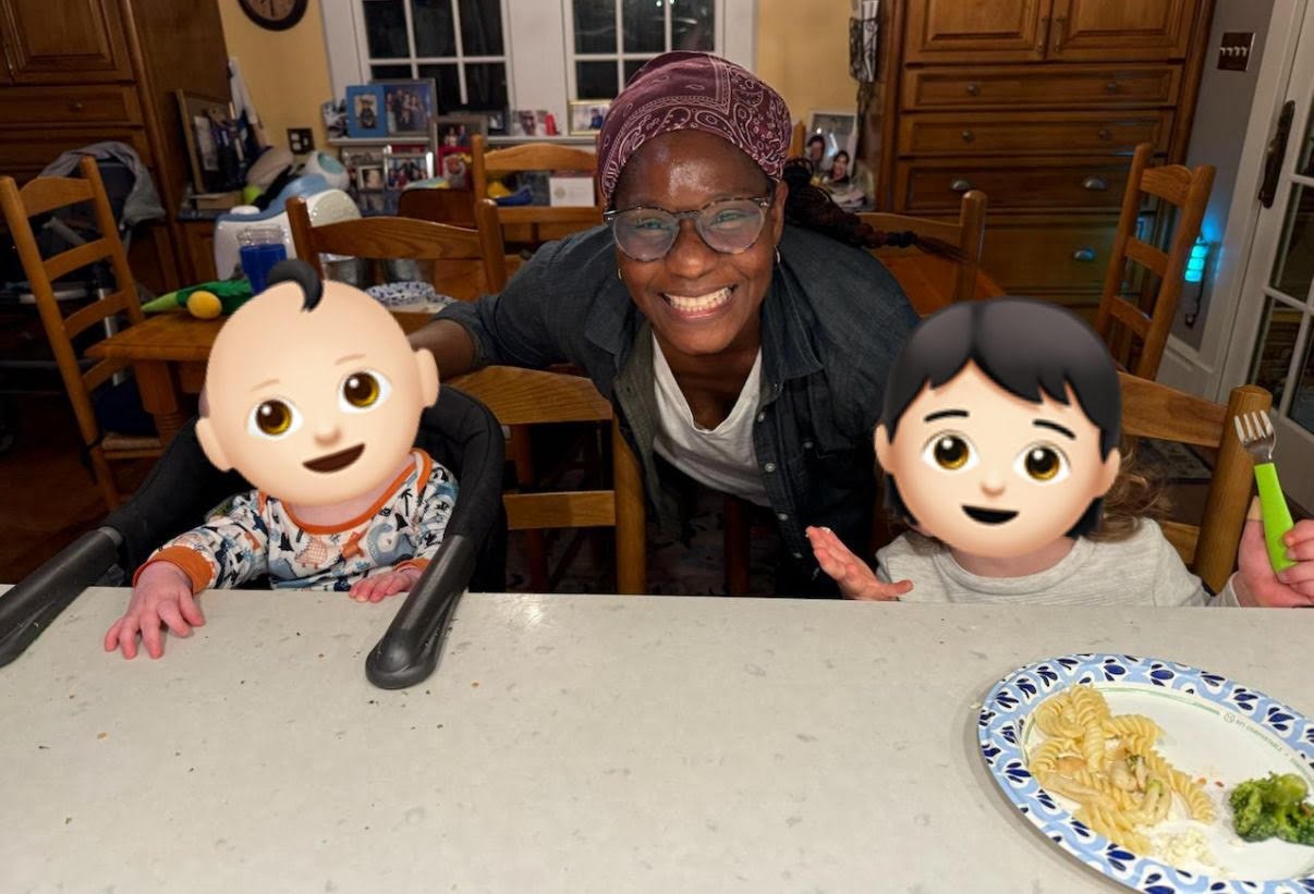 A woman smiling at a table with two children, one of whom is in a high chair. The children's faces are covered with animated emoji masks. There is a plate of food on the table, and the background shows a kitchen with wooden furniture and various items.