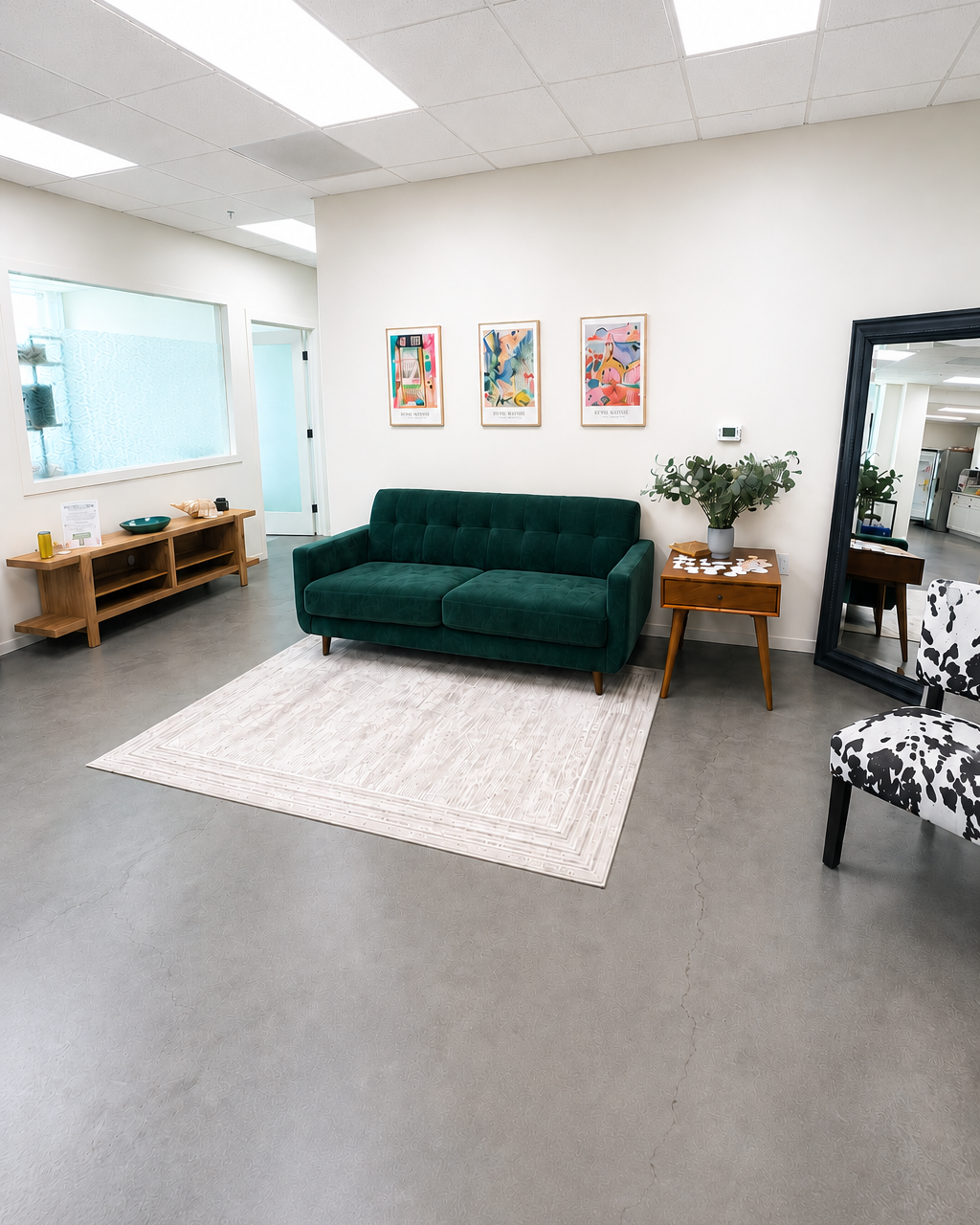 Modern waiting room with a green sofa, a small wooden side table with a plant, framed artwork on the wall, a large mirror, and a patterned accent chair.