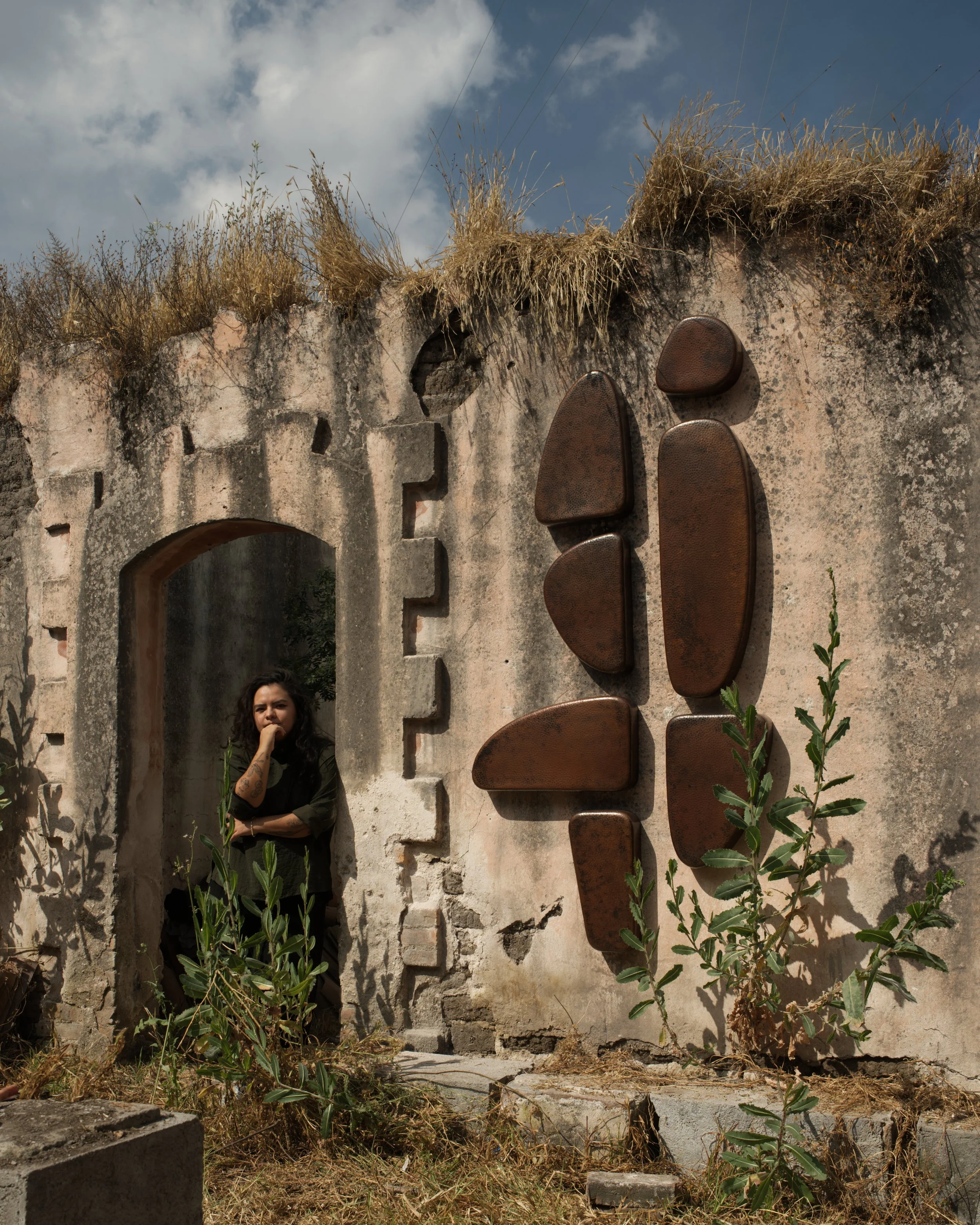 Mujer con cabello rizado de cabello oscuro, vistiendo ropa oscura, de pie en una abertura en un muro deteriorado con esculturas de hierro en la pared, plantas creciendo en el suelo y un cielo parcialmente nublado en el fondo.