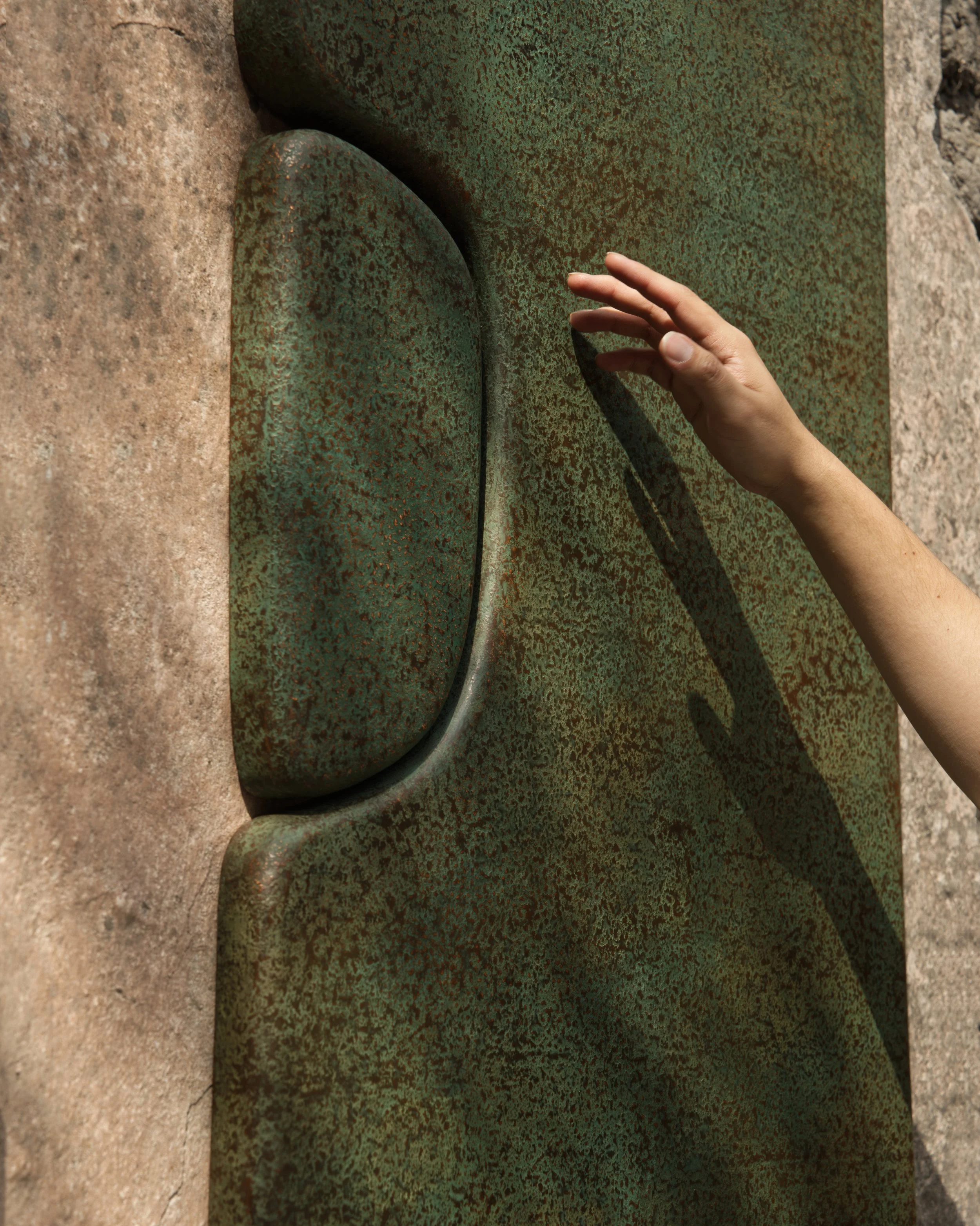 Una mano tocando una escultura de piedra con acabado verde oxidado y textura áspera, ubicada en una superficie de piedra.