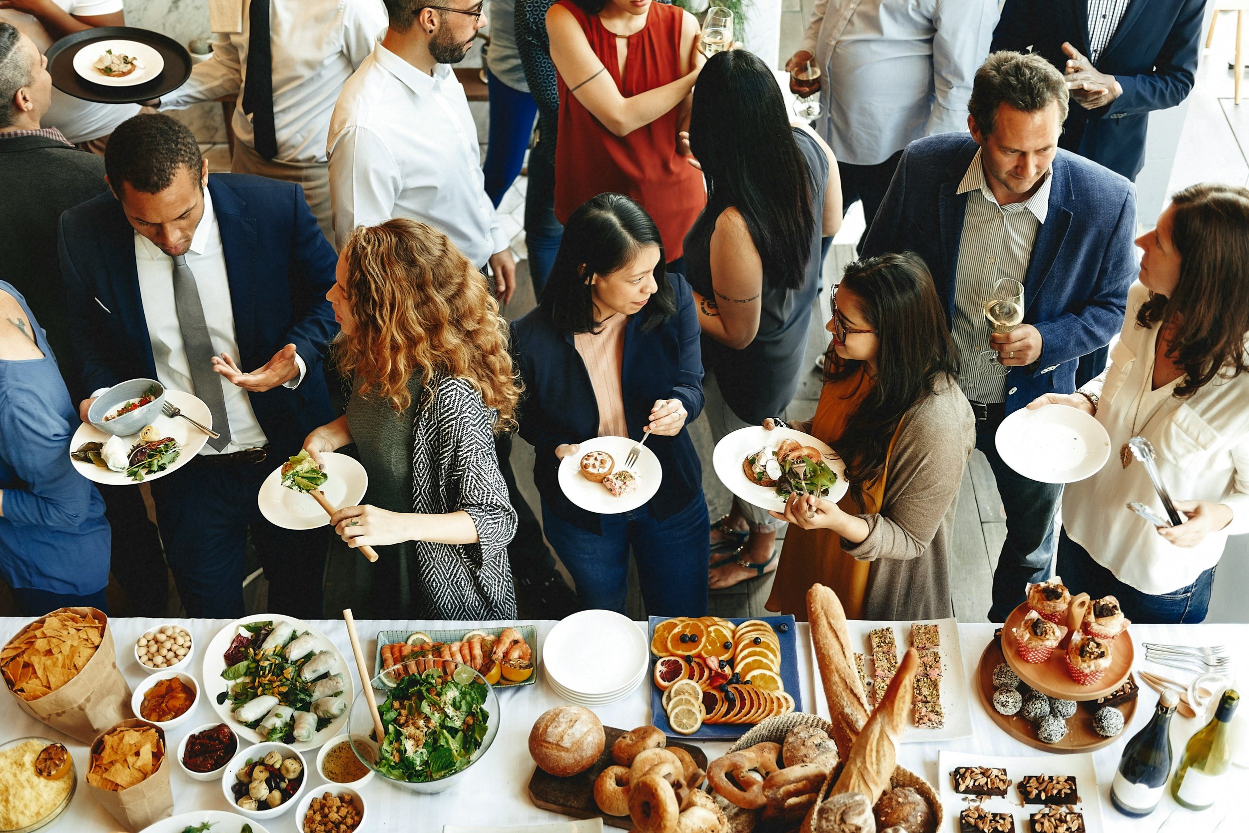 People at a social gathering or party selecting food from a buffet with various dishes, drinks, and desserts on the table.