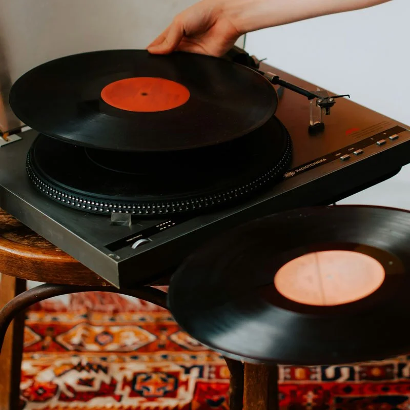 A person handling a vinyl record on a turntable with two records, one on the turntable and one on a nearby table, in a room with a patterned rug.