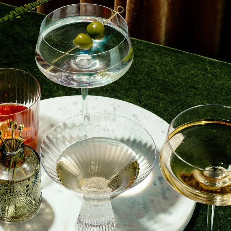Different cocktail glasses on a marble table with olives in a martini glass, a red drink in a glass, and a dark drink in another glass.