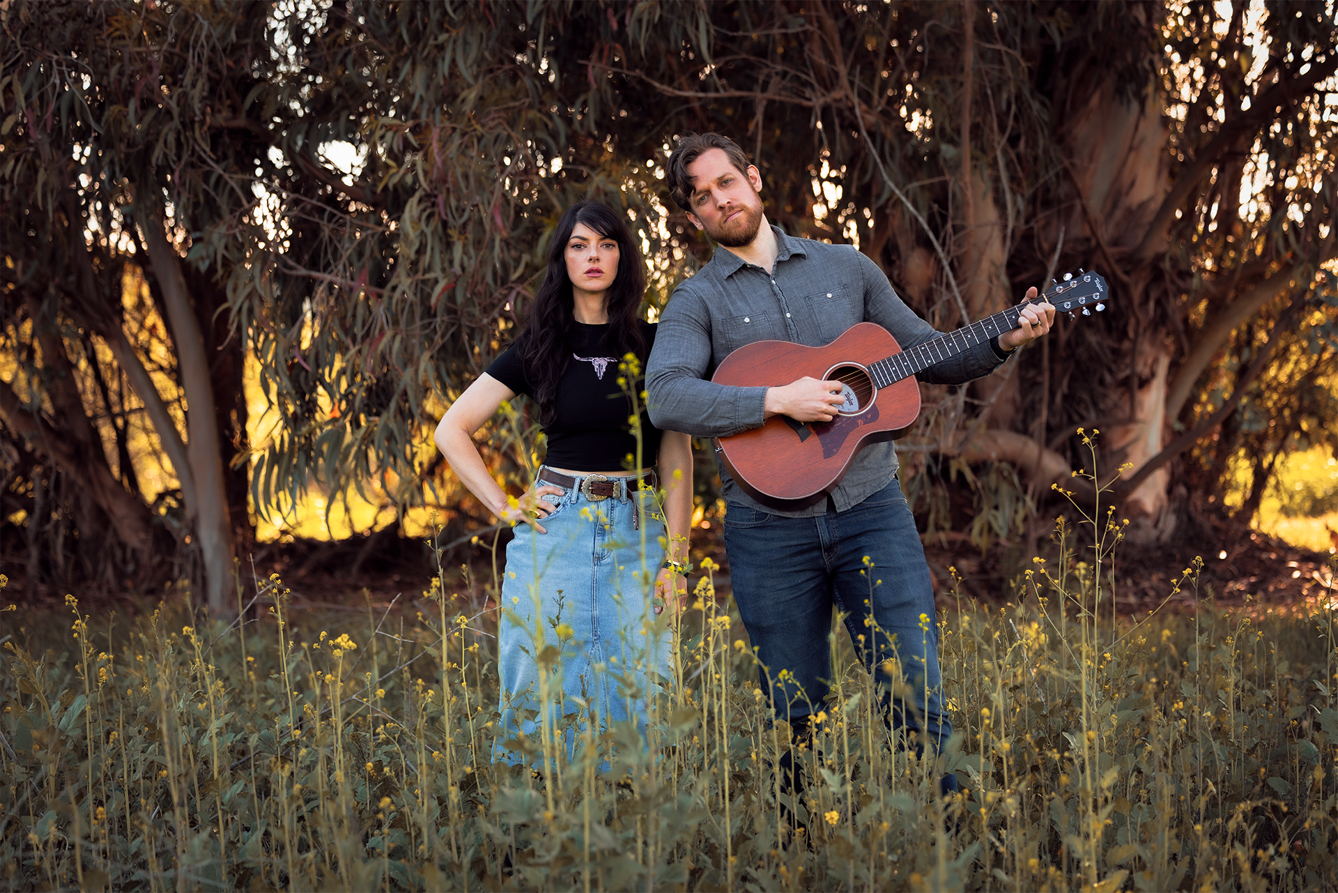 A man playing acoustic guitar and a woman standing next to him in a field near a large tree during sunset.