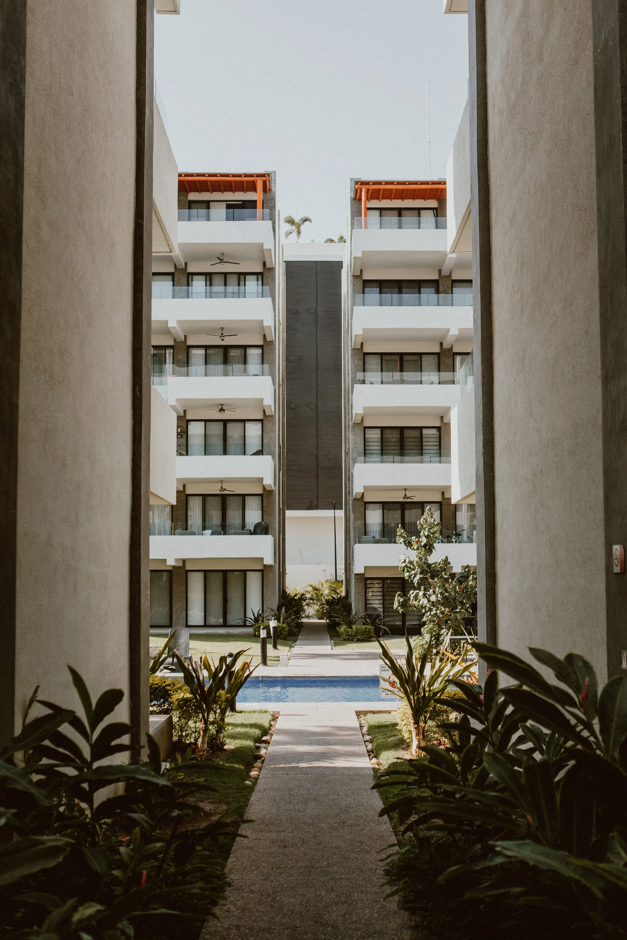 View of a modern apartment complex from between two buildings, with a pathway leading to a swimming pool and garden area.