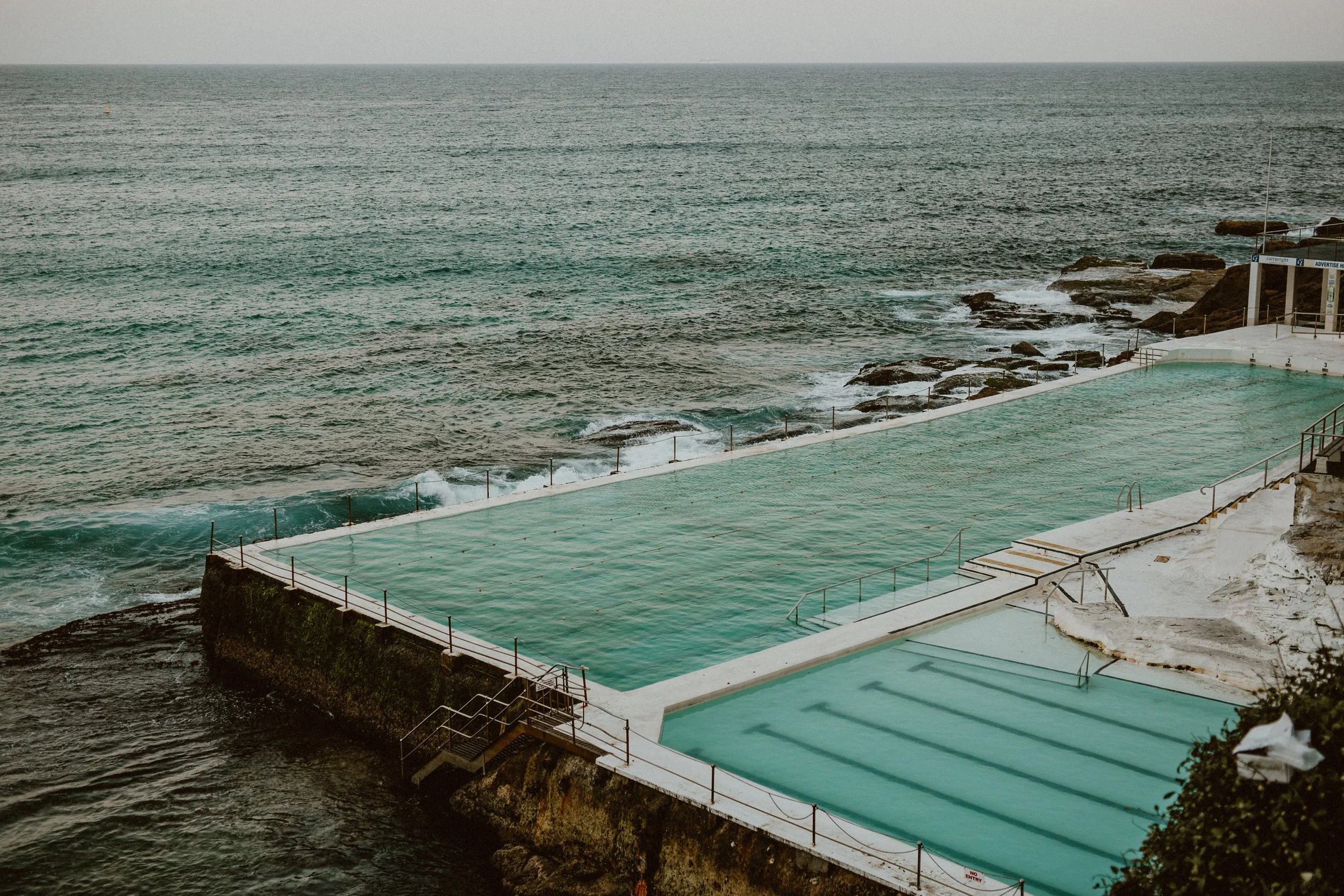 Coastal swimming pools built on rocks overlooking the ocean, with a rocky shoreline and calm sea in the background.