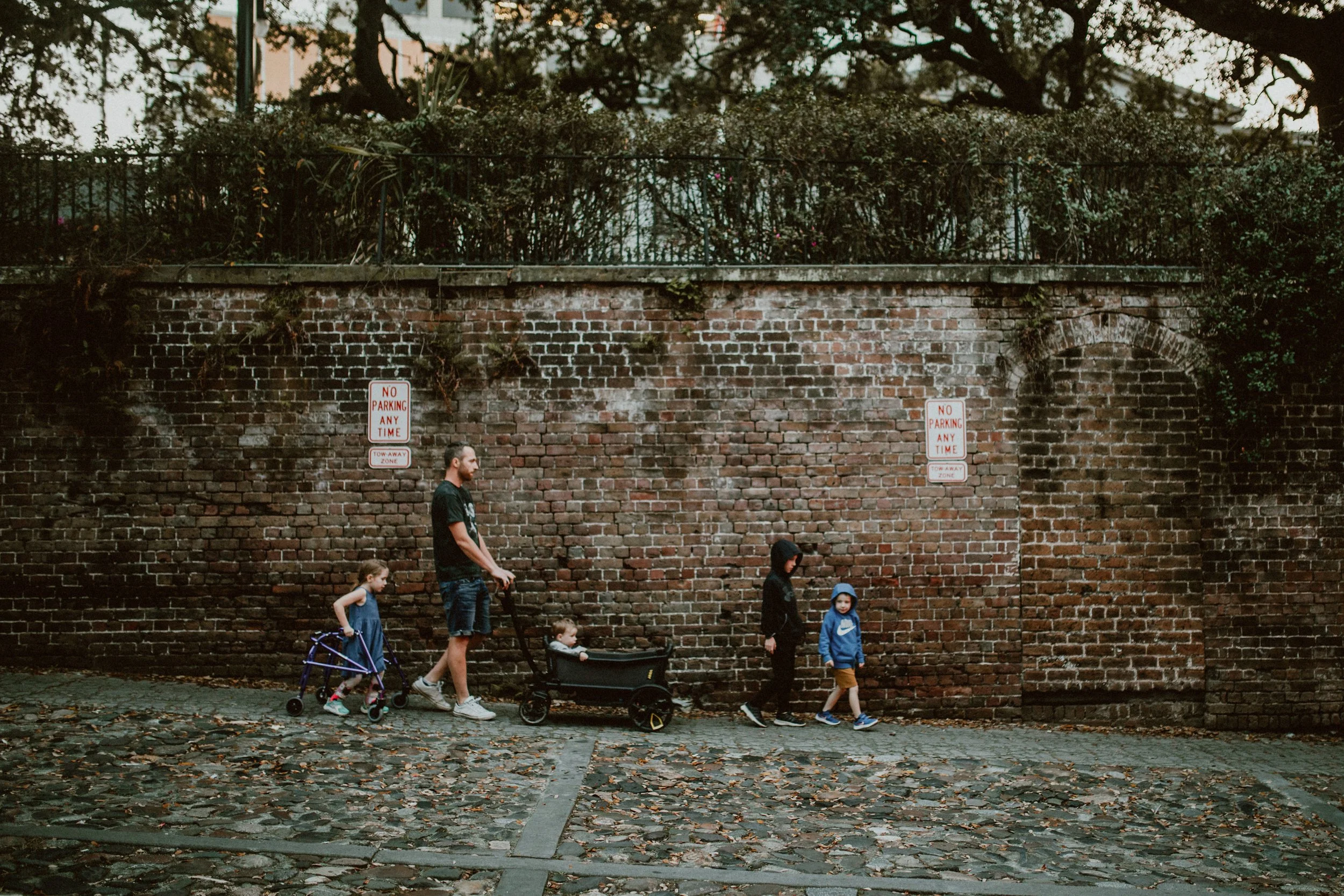 A group of five people, including two children and their parents, walk along a brick wall sidewalk with fallen leaves in the foreground. The man pushes a stroller, while the woman walks with a young girl and a boy. The scene is outdoors in an urban a