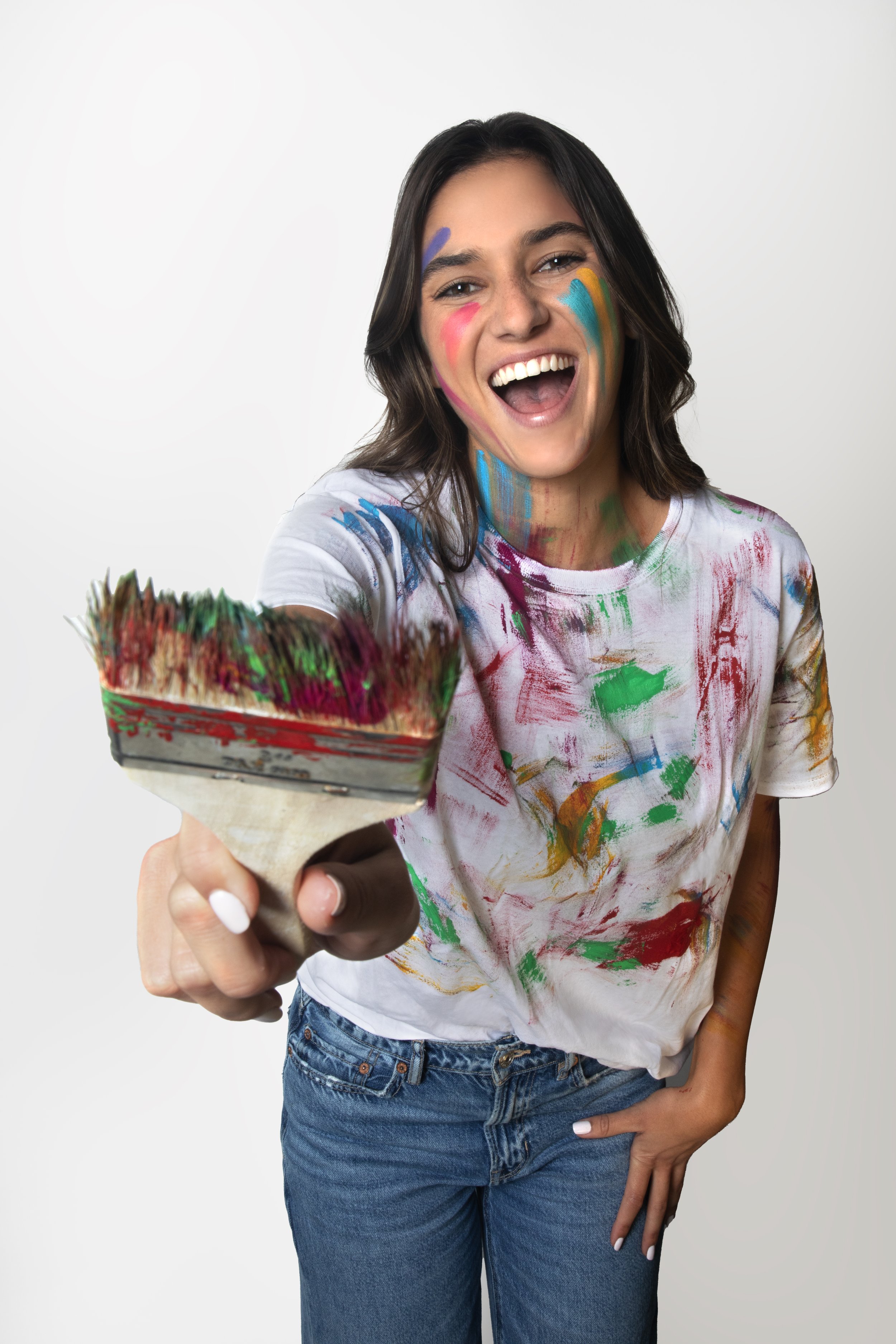Person smiling with paint-splattered shirt and face, holding a paintbrush.