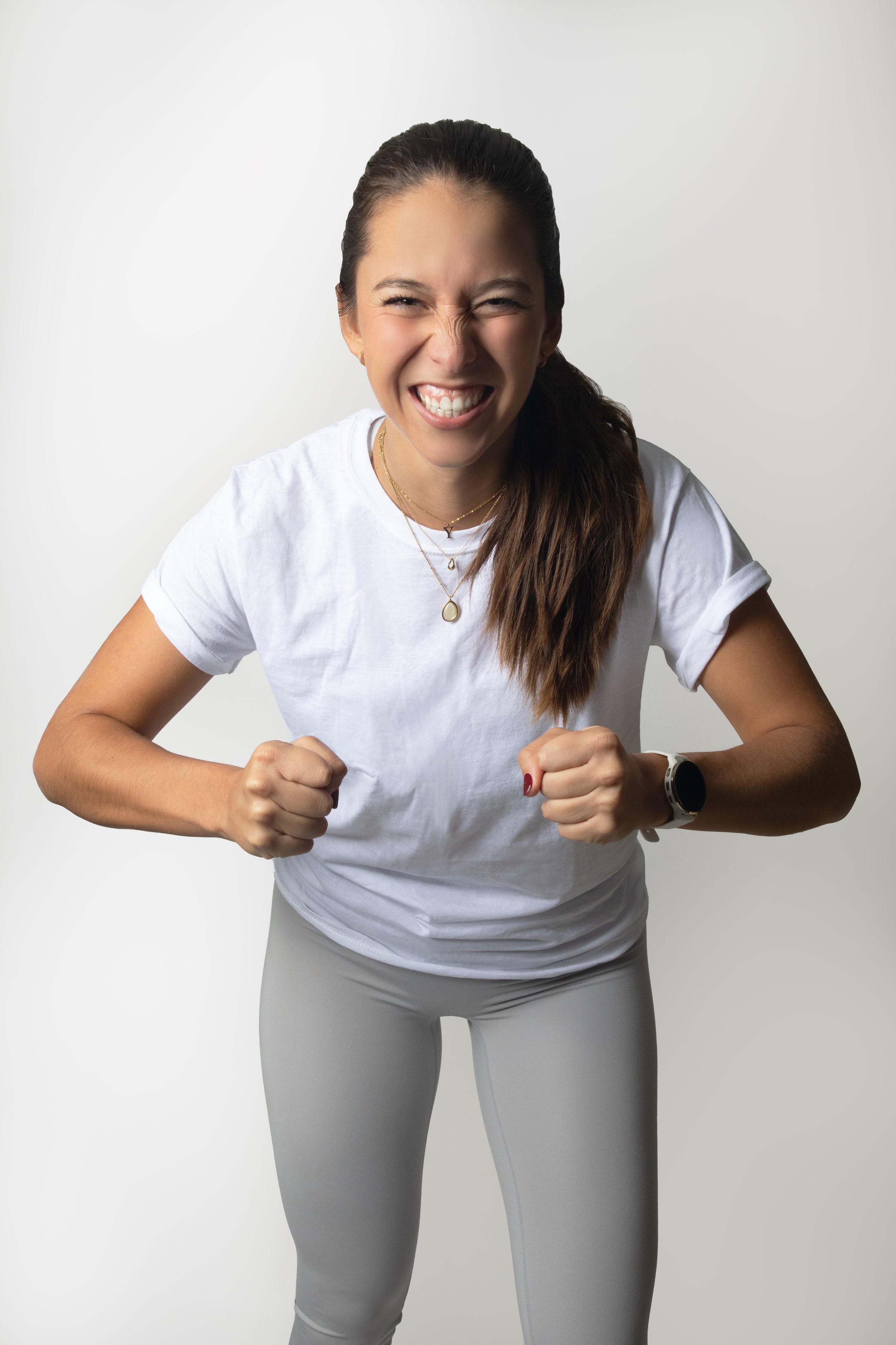 Young woman in white T-shirt and gray leggings posing energetically with a big smile, against a plain background.