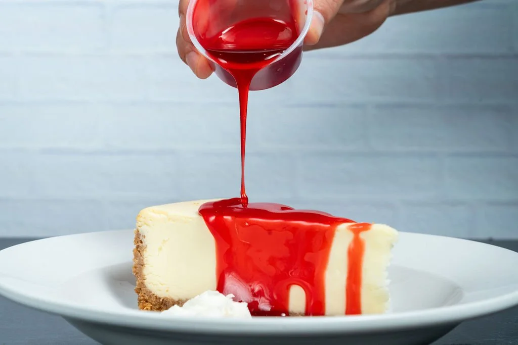 A slice of cheesecake on a white plate with red fruit sauce being poured over it from a glass, with a small dollop of whipped cream on the side.